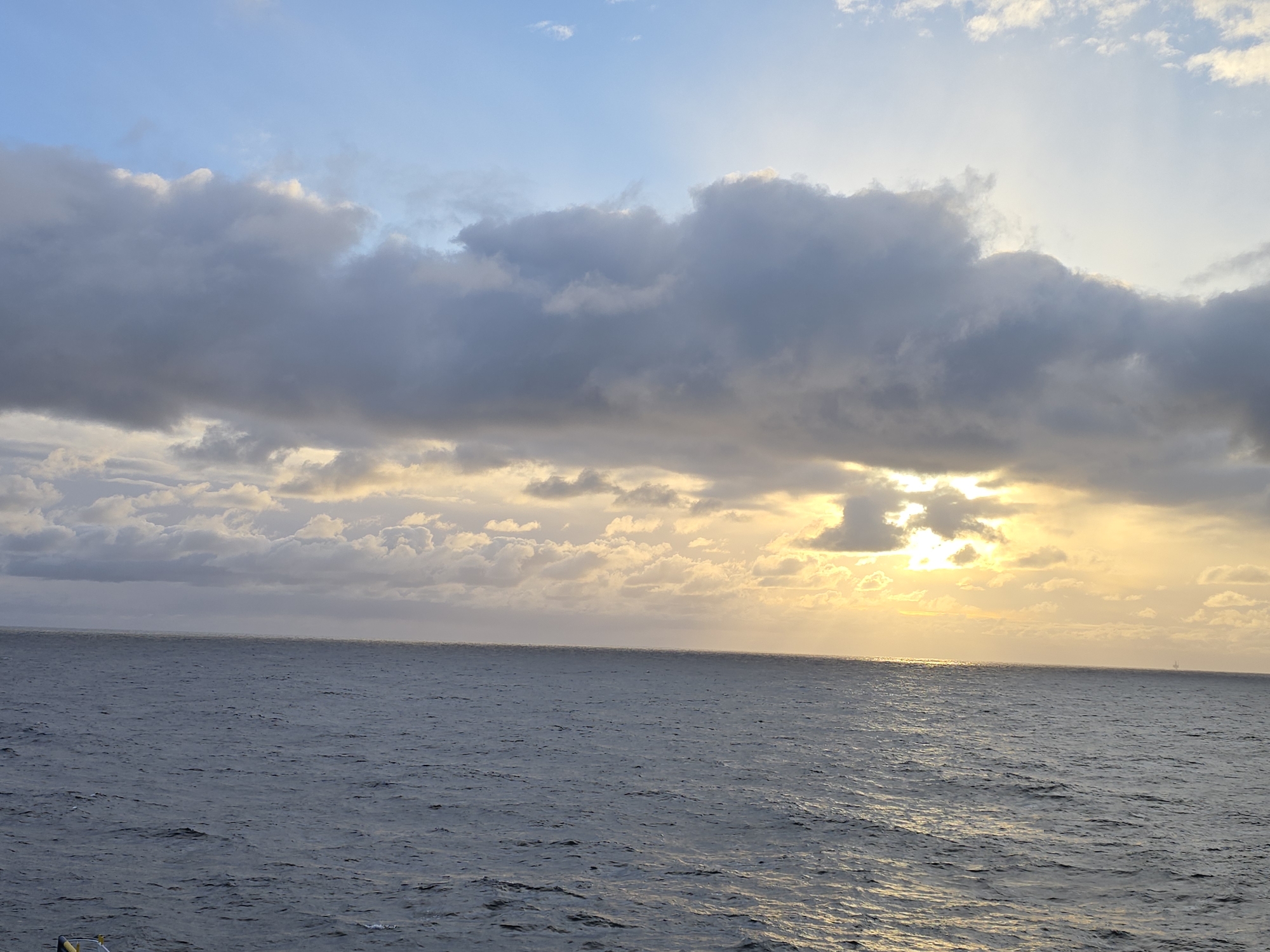 The image shows an ocean scene under a cloudy sky with the sun partially obscured, creating a warm glow on the horizon and reflecting on the water's surface. The location is Unknown, Unknown. There are no people present. A distant structure, possibly an offshore platform, is visible on the horizon.