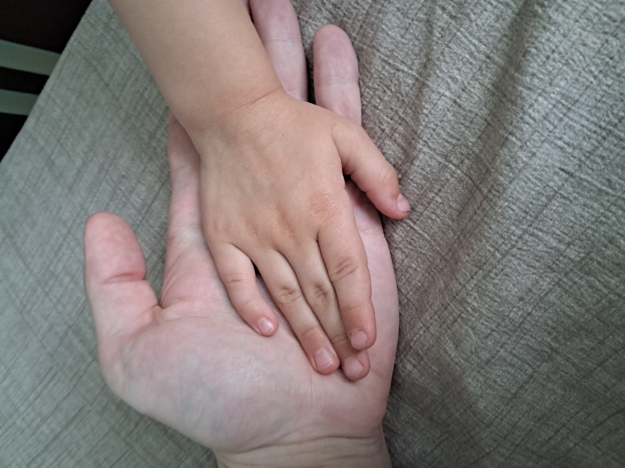 Two hands are in the foreground, one large adult hand and one small child's hand. The child's hand is resting on top of the adult's hand, with the fingers of the child's hand extended across the palm of the adult's hand. The background is a textured, woven fabric, likely a bedspread or tablecloth, in a neutral beige or light brown color. The lighting is soft and diffused. The image is a close-up shot, focusing entirely on the hands and the immediate background. There are no visible landmarks or environmental cues to indicate a specific city or location.