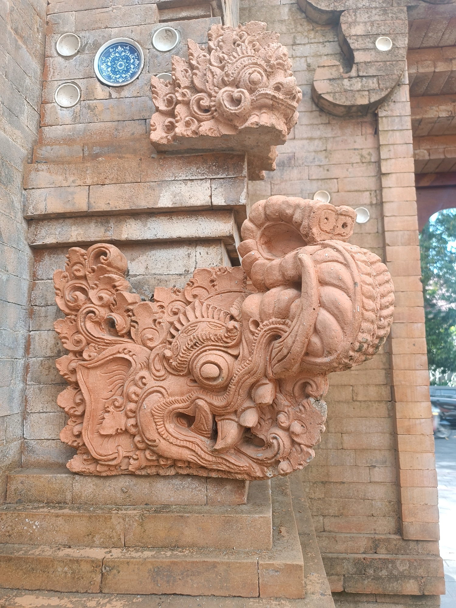 An ornate, terracotta-colored dragon head sculpture is mounted on a brick wall near an archway. The sculpture has intricate details, including large eyes, elaborate horns, and a gaping mouth. Above it, a smaller, similar dragon head sculpture is also visible. The brickwork of the wall is a reddish-brown hue, and the wall is adorned with several circular white ceramic decorative elements and one blue and white patterned ceramic plate. The background shows an archway and foliage, suggesting an outdoor setting, likely in the city of Tabanan, Indonesia, given the architectural style. No people are present in the image.