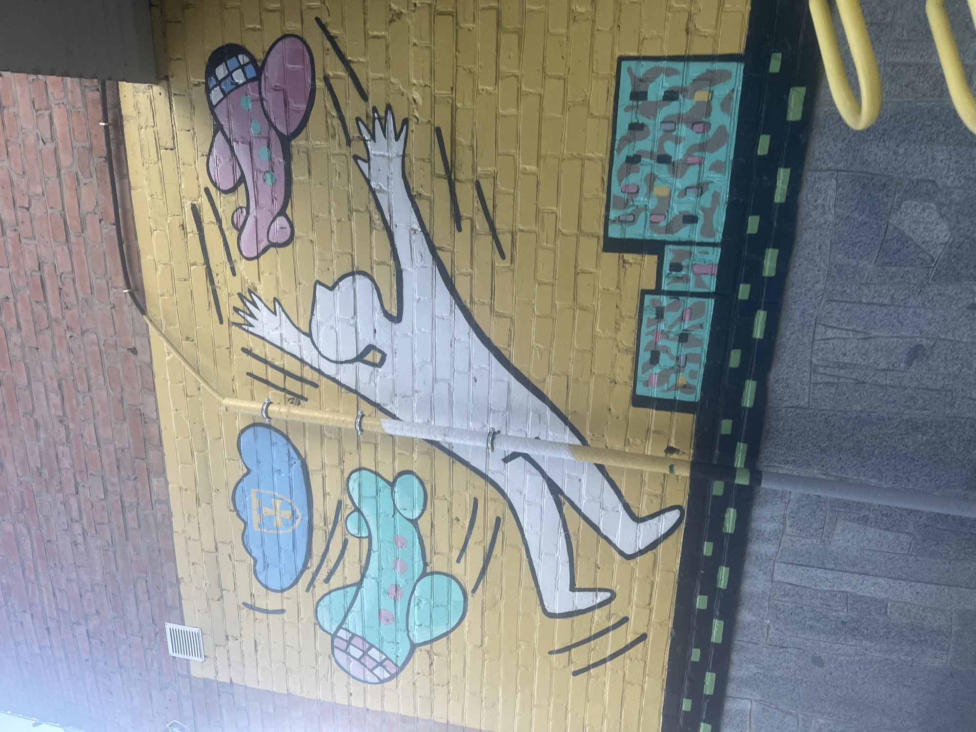 The image displays a mural painted on a yellow brick wall. The mural features a stylized white humanoid figure with arms outstretched, appearing to be in mid-air. Two cartoonish airplanes are depicted flying around the figure: one light blue with pink polka dots to the left, and one pink with light blue polka dots to the right. Above the figure, a blue cloud with a yellow emblem is visible. The bottom of the mural depicts a dark band with green squares, resembling a road or ground. The wall is topped by a section of red brick. A metal ventilation grate is present on the upper left portion of the yellow wall. A yellow pole, possibly a lamppost or part of a structure, runs vertically through the mural, partially obscuring the white figure. The ground below the mural is made of gray stone slabs. This mural is located in the city of Horodok, Ukraine.