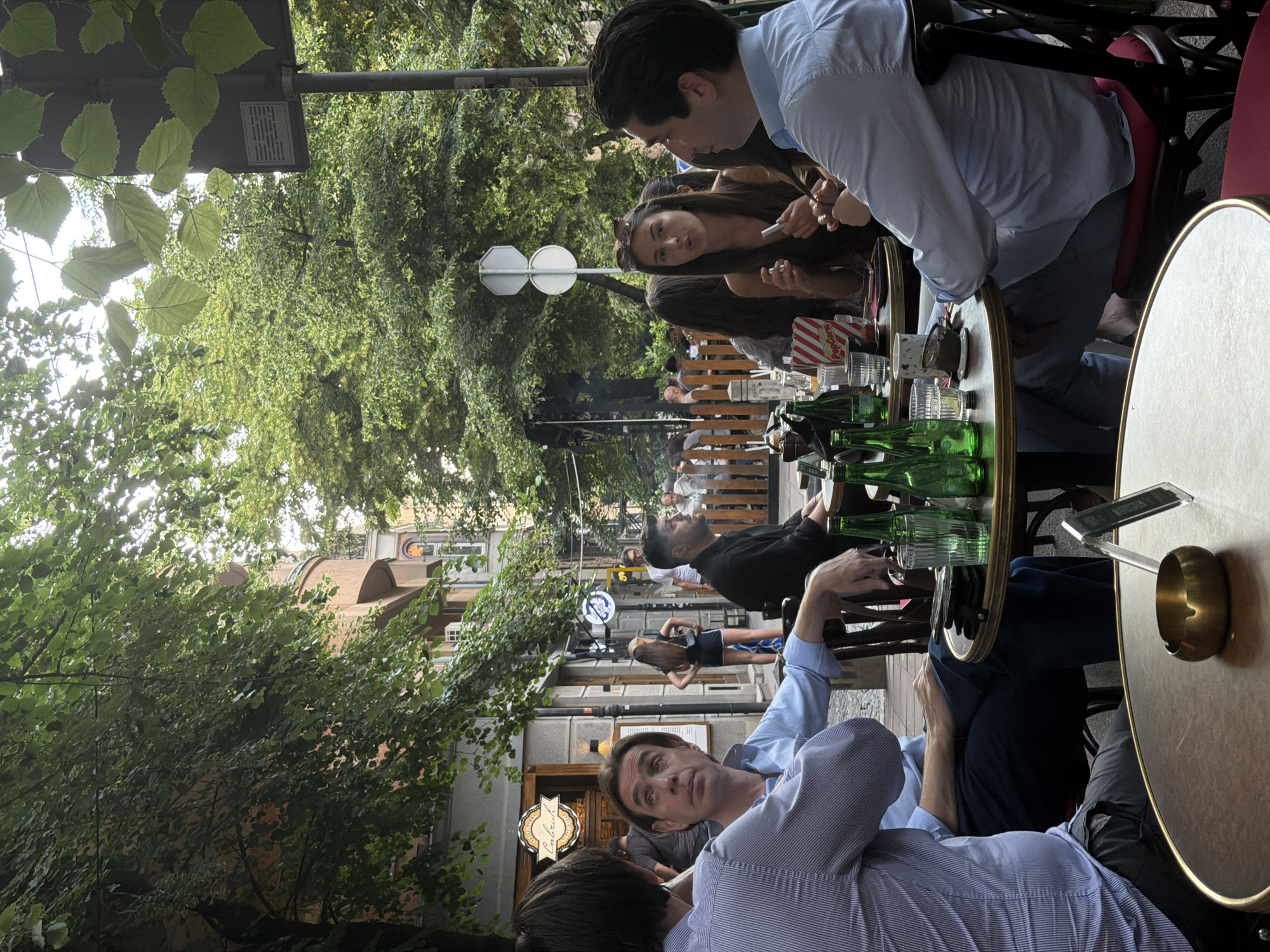 The image depicts an outdoor cafe scene in Belgrade, Serbia. Several people are seated at tables. There is a man in a light blue shirt leaning over a table, engaged in conversation with someone out of frame. At another table, a man in a black shirt and another man in a light blue shirt are seated. A woman with long brown hair is also seated at a table with a man in a light blue shirt. Green glass bottles and glasses are on the tables. In the background, other people are walking on the sidewalk. Trees line the street, and buildings are visible. A street sign can be seen.