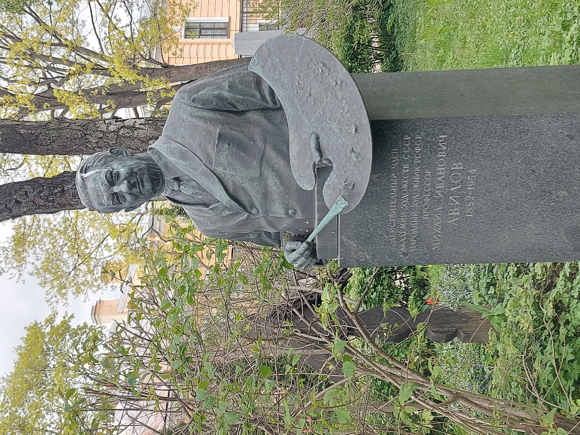 The image shows a bronze monument to the Soviet painter Mikhail Ivanovich Avilov. He is depicted in a suit with a palette and brush in hand. The pedestal of the monument contains text in Russian.
The monument is located in Saint Petersburg, Russia. The sculpture is surrounded by greenery. A building is visible in the background.