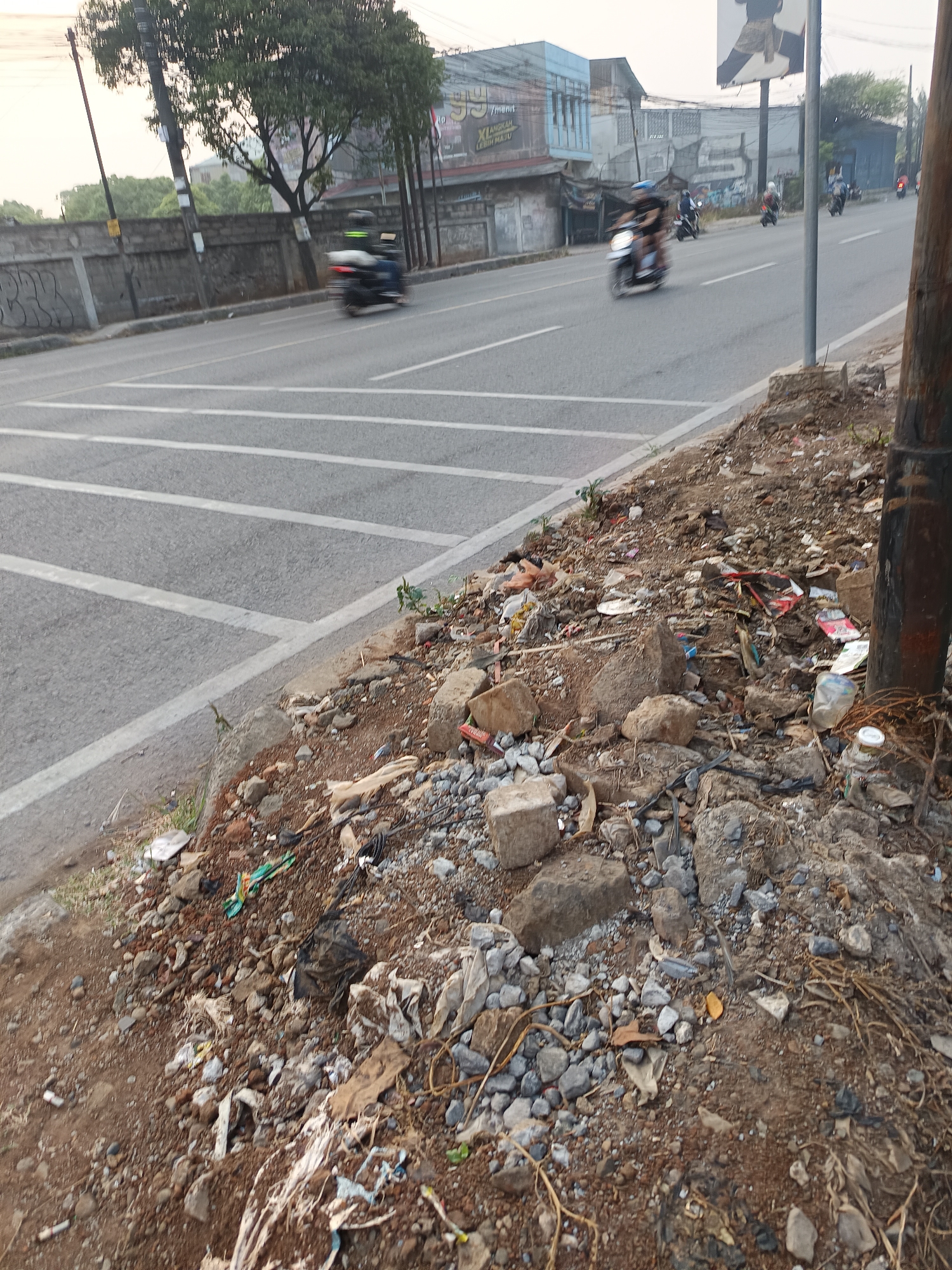 The image depicts a roadside scene in Talaga Sari, Indonesia. A paved road with white lane markings runs horizontally across the image, with several motorcycles visible in motion on it. To the right of the road is a dirt embankment covered in rocks and scattered debris, including plastic bottles, wrappers, and other trash. A wooden utility pole stands on the embankment, its base surrounded by debris. In the background, buildings and trees line the opposite side of the road. The sky is overcast.