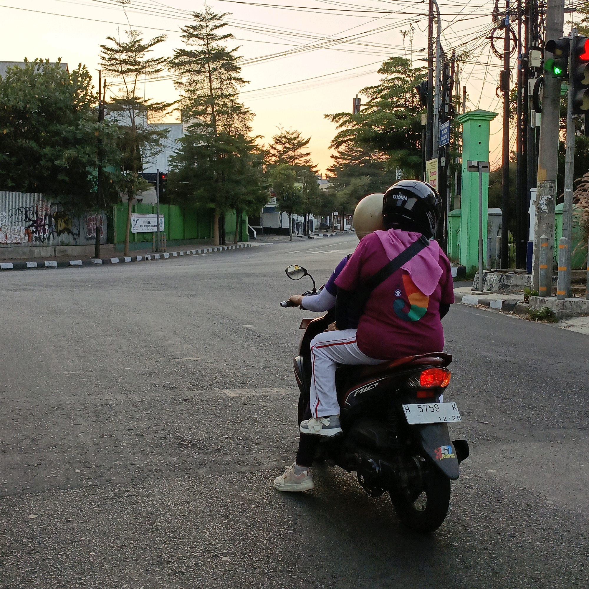 Two individuals are riding a motorcycle down a paved road in Salatiga, Indonesia, during a sunset. The person in the back is wearing a maroon shirt with a colorful design on it, white pants with red stripes, and light-colored sneakers. They are wearing a black helmet with "YAMAHA" written on it. The person in front is wearing a beige helmet and a dark purple shirt. The motorcycle is black and has a license plate that reads "H 5759 K" with "12.28" below it. On the left side of the road, there is a metal fence covered in graffiti and some trees. To the right, there are traffic lights, utility poles with many wires, and a tall green pillar. The sky is a soft orange and yellow, indicating the sun is setting.