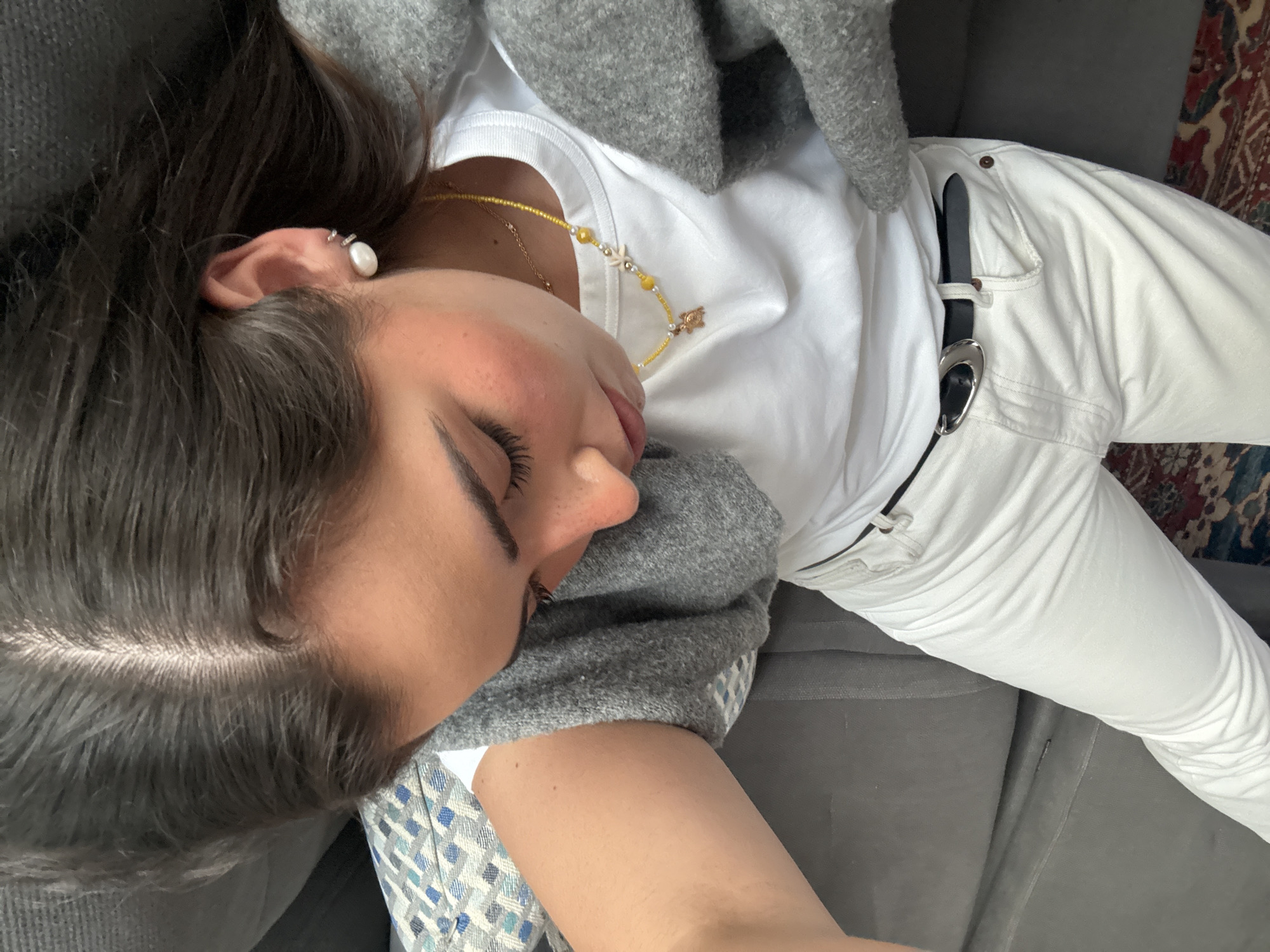 A woman is lying on a gray couch. She is wearing white pants, a white t-shirt, and a gray sweater. She has dark hair and is wearing a pearl earring and a beaded necklace. The location is unknown and there is no evidence to suggest it is in San Bernardino Tlaxcalancingo, Mexico.