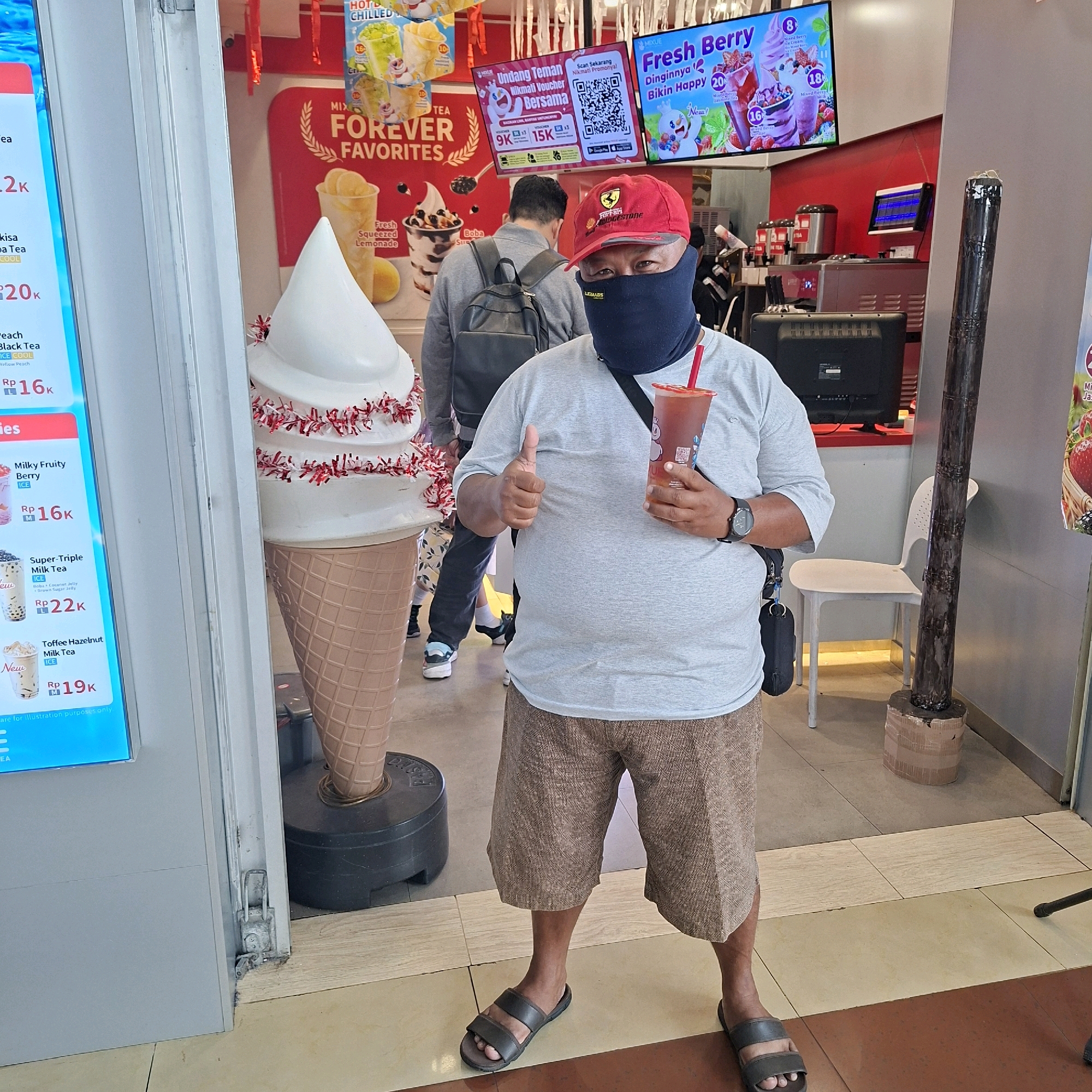 In an indoor setting, a man wearing a grey t-shirt, brown patterned shorts, a red baseball cap, and a black face mask stands holding a drink with a red straw and giving a thumbs-up. He is positioned in front of a large decorative ice cream cone and a menu board. Behind him, other people are visible, and a counter area with beverage machines is present. The environment appears to be a food establishment, possibly a cafe or drink shop.