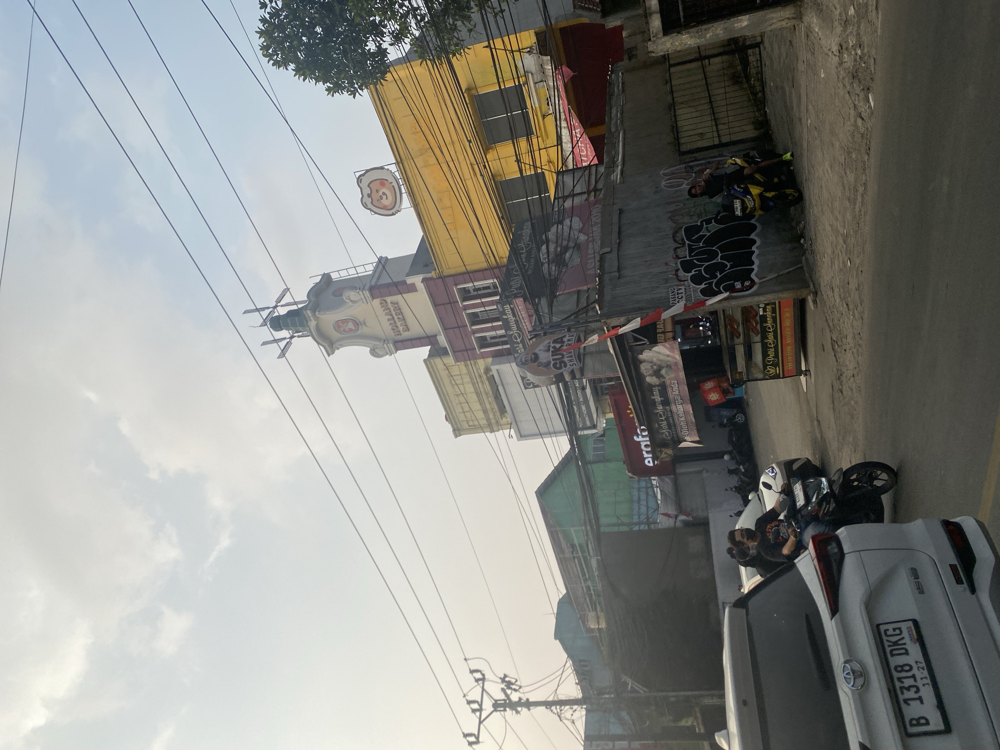 This image captures a street scene in Depok, Indonesia, likely during daylight with overcast skies. The foreground features the rear of a white Toyota SUV, displaying a license plate that reads "B 1318 DKG". Next to it, a man wearing a black t-shirt and a face mask rides a black motorcycle. Behind them, a yellow building is prominent, topped with a sign featuring a cartoon bear. A taller, multi-story building with a tower-like structure and the words "HOLLAND BAKERY" is partially visible to the left of the yellow building.
Further down the street, several other buildings of varying colors and designs are visible, including a green-painted structure. Overhead, a dense network of power lines crisscrosses the sky, supported by utility poles. The street itself appears to be paved. Several other motorcycles are parked or in motion along the roadside, and graffiti adorns a concrete wall in the middle ground. A food stall or small shop with signage is also present on the right side of the image. The overall impression is of a busy urban environment with commercial establishments and active street life.