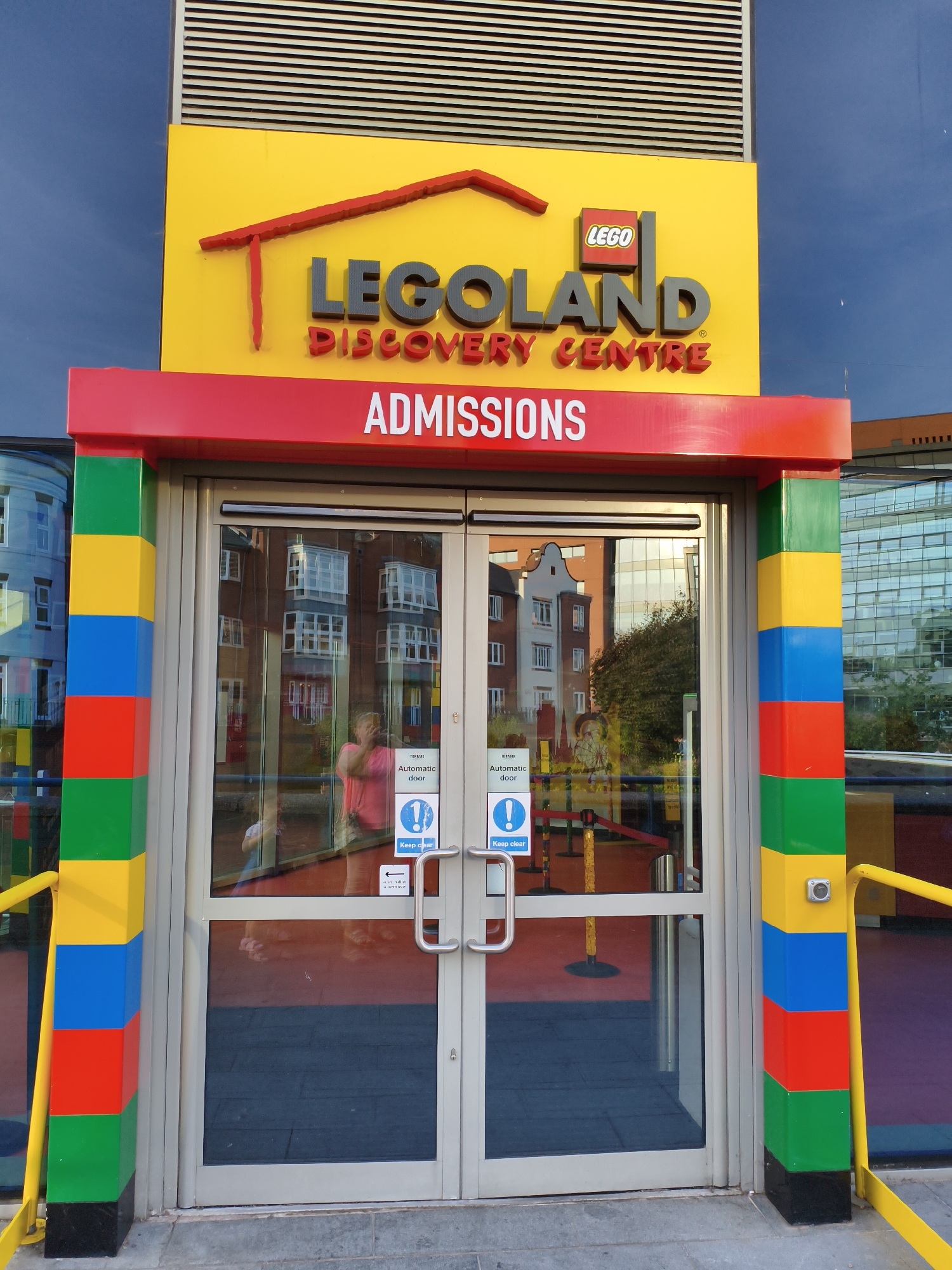 The image shows the entrance to the Legoland Discovery Centre in Birmingham, United Kingdom. The entrance is brightly decorated with the Lego brand colors. Above the entrance, a yellow sign displays the Lego logo and the words "LEGOLAND DISCOVERY CENTRE" in large grey letters. Below this, a red sign reads "ADMISSIONS" in white letters. The entrance itself consists of double glass doors, with colorful Lego-like pillars (red, blue, yellow, and green) on either side. Inside the building, reflections in the glass show people and the interior of the attraction. A woman in a pink shirt is visible in the reflection of the right door, holding a phone. Signs on the doors indicate they are automatic and to "Keep clear." Yellow railings are also visible, leading away from the entrance. The overall environment appears to be an outdoor public space, with a blue sky overhead and other buildings reflected in the glass.