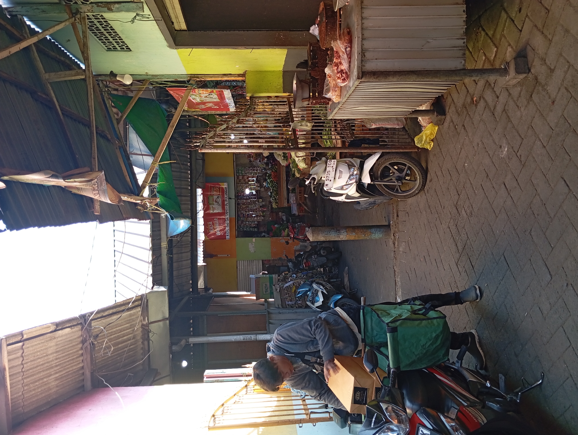 A man is seen in the foreground, hunched over a cardboard box on the seat of a motorcycle. He is wearing a grey hoodie, dark pants, and dark sneakers. The motorcycle, a red and black Honda, has a large green bag attached to its rear. In the background, a market scene unfolds. Stalls are set up under corrugated metal roofs, with various goods displayed, including produce and what appears to be raw meat on a counter. Several other motorcycles are parked throughout the market. The environment suggests a street market in Kepulungan, Indonesia, with the architecture and general atmosphere indicative of the region. The lighting suggests it is daytime, with sunlight casting shadows.