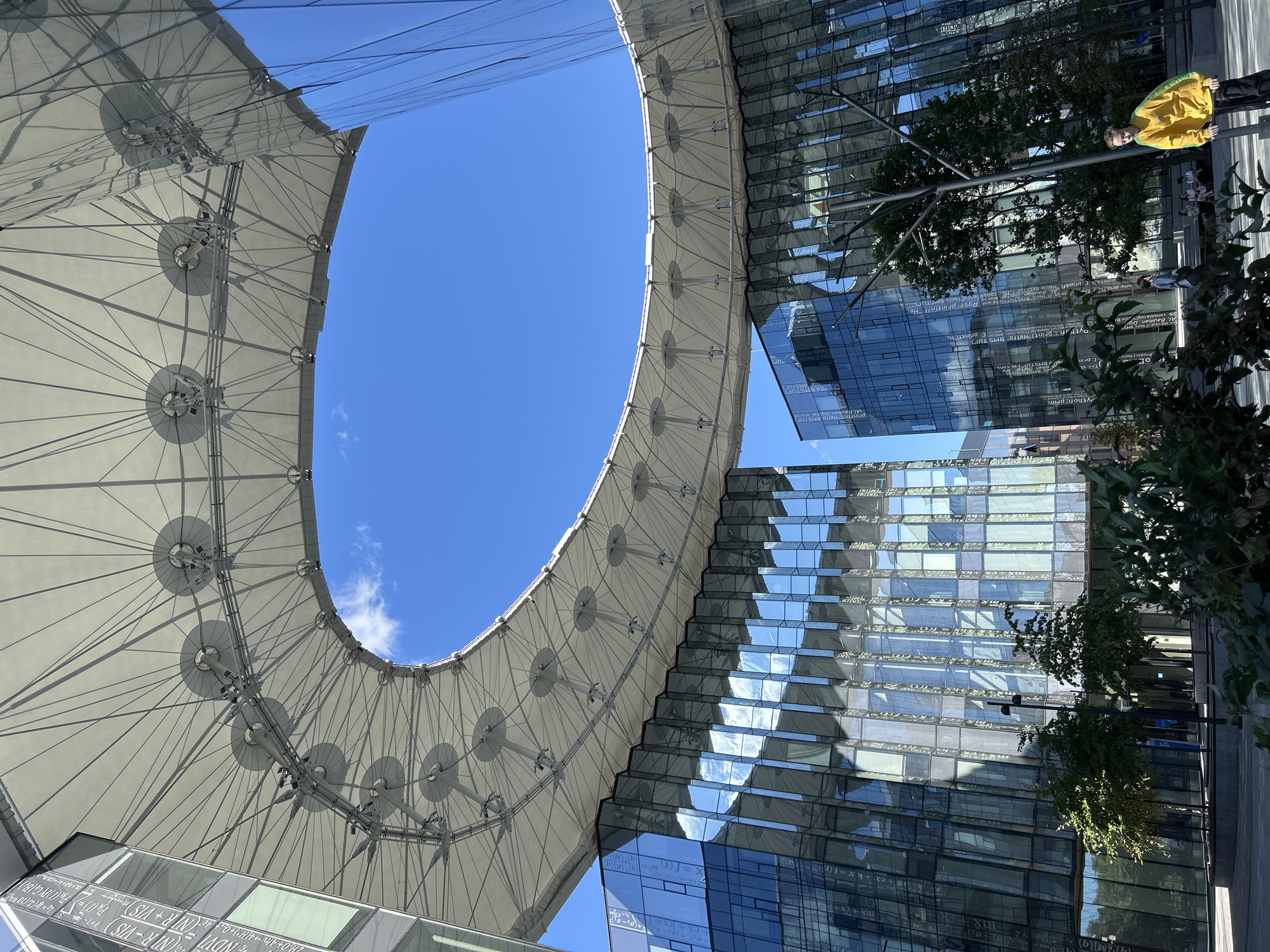A young boy in a yellow sweatshirt and black pants stands in front of a modern glass building on a sunny day. The boy is holding a scooter, and his gaze is directed towards the camera. In the background, the glass facade of the building reflects the sky and surrounding trees. An overhead canopy structure, supported by cables, is visible above the boy and the building. Several people are seated on a bench in the distance, and trees are planted around the base of the building.