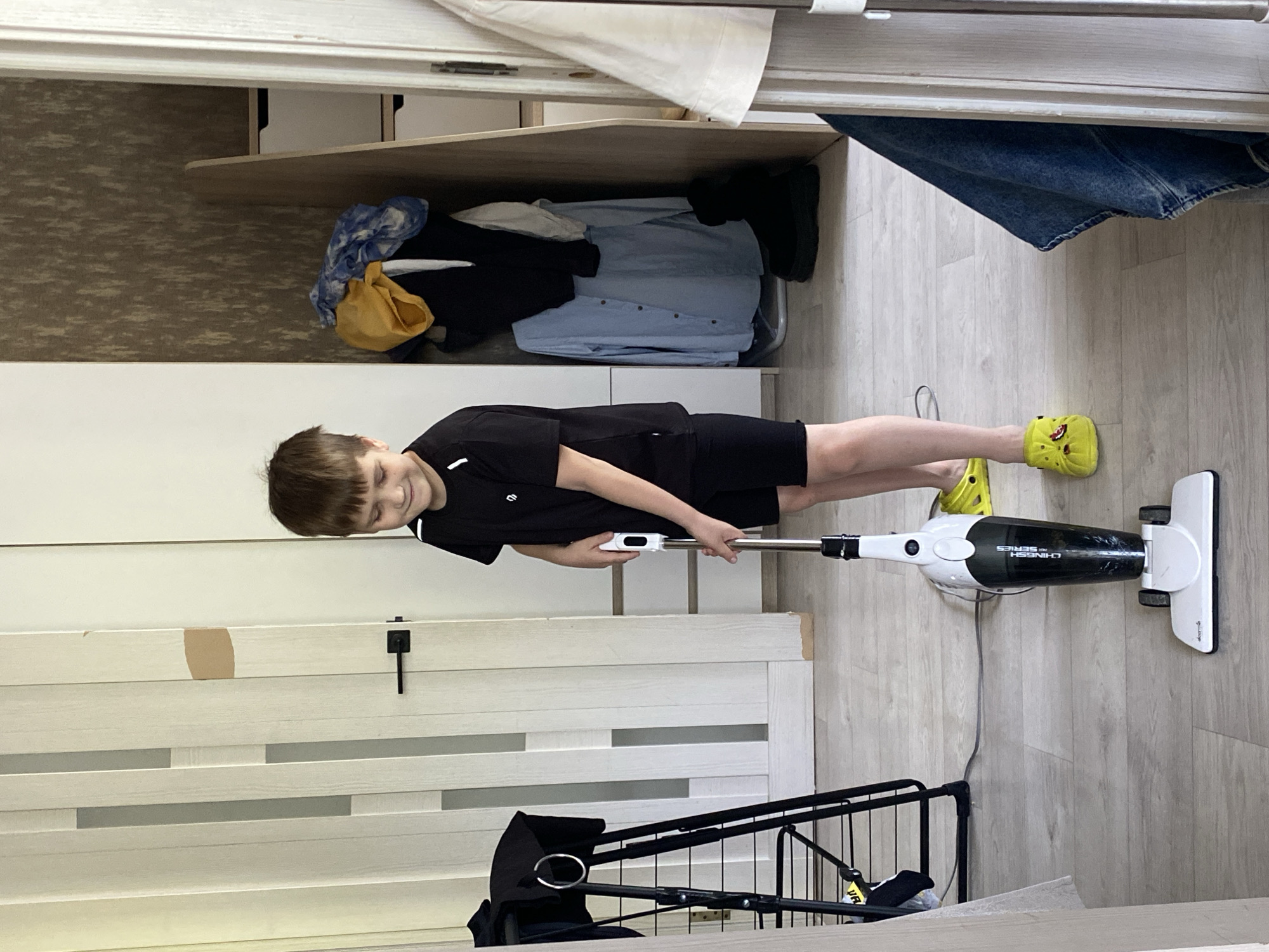A smiling boy, wearing a black t-shirt and shorts, operates a black and white Deerma vacuum cleaner on a light-colored wooden floor. He wears yellow Crocs. The scene takes place inside a room, likely a home in Chaikovsky, Russia. To the left is a multi-panel white door with a dark handle, and behind the boy is a closet with shelves containing clothes. A black stroller frame is visible in the lower left corner.
