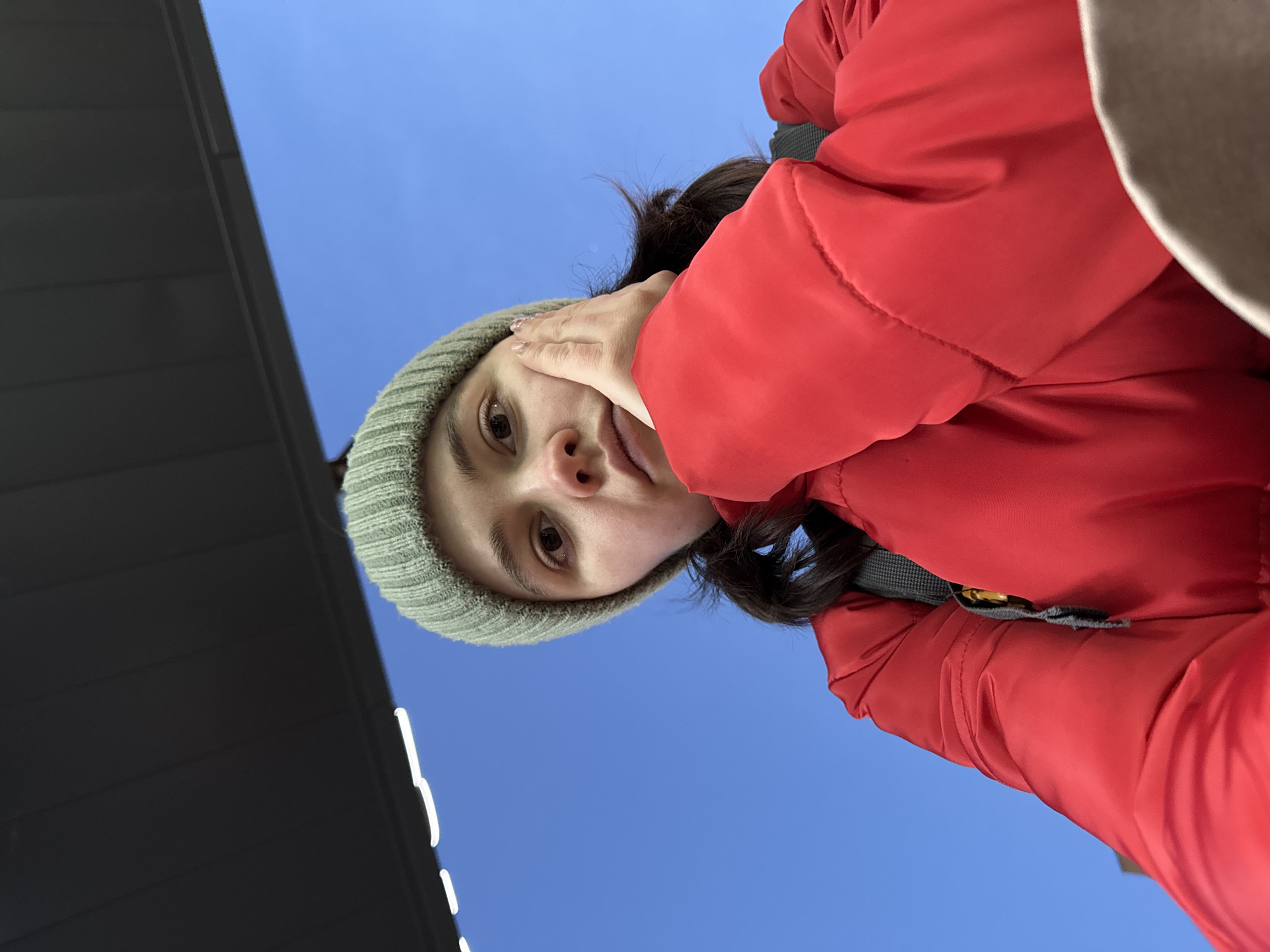 A woman wearing a grey beanie and a red puffer jacket is looking down at the camera. Her right hand is touching her face. The background shows a blue sky and a dark grey building. There is no specific information in the image to confirm that the location is in Izhevsk, Russia.