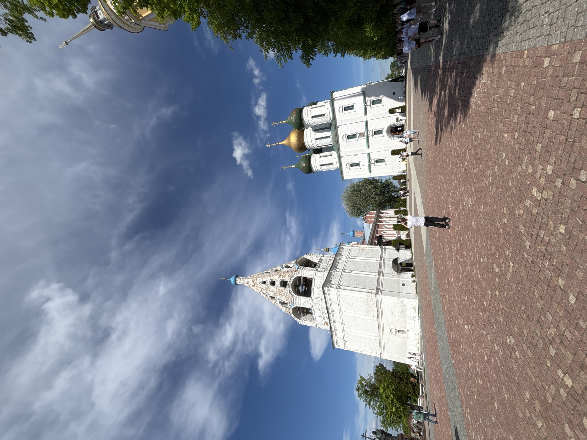 The image captures a view of Cathedral Square in Kolomna, Russia. Dominating the frame are two prominent architectural structures: the five-domed Dormition Cathedral with its golden and green domes, and the bell tower of John the Baptist. Both buildings are white with arched window detailing. The sky is a bright blue with scattered white clouds. The foreground features a cobblestone paved square with a path of darker colored stone in the middle, populated with people. The surrounding area is lined with green trees.