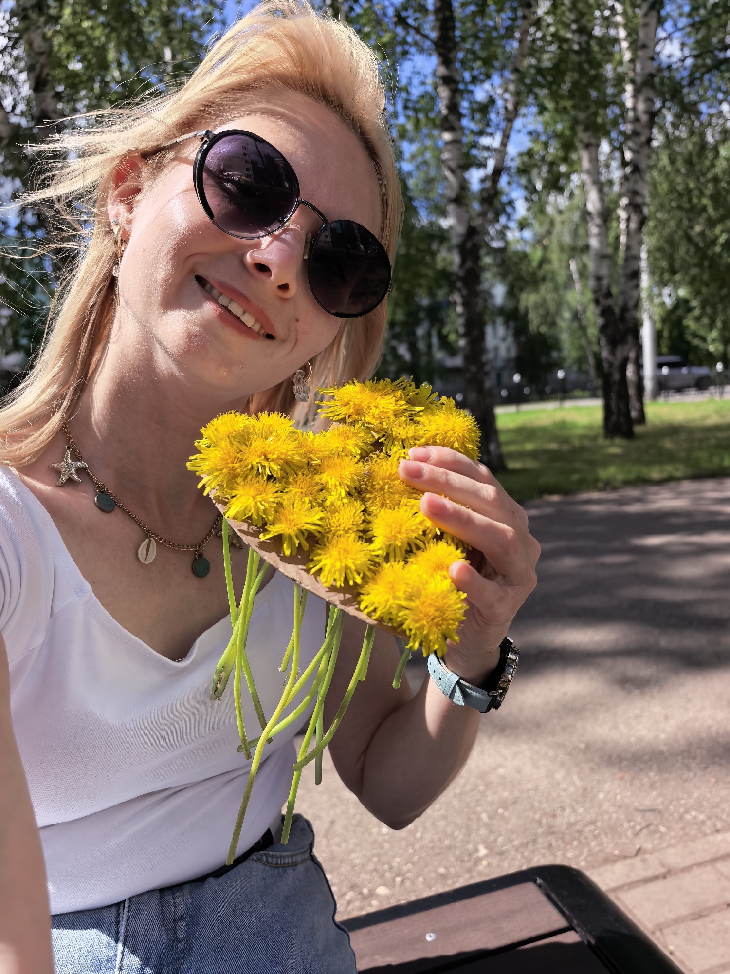A fair-skinned woman with blonde hair and sunglasses holds a bouquet of dandelions. She is wearing a white top, denim shorts, and a necklace with seashell and coin charms. A watch with a light blue band is visible on her wrist. The background shows trees and a park path, suggesting an outdoor setting in Sterlitamak, Russia. The woman appears to be smiling and taking a selfie.