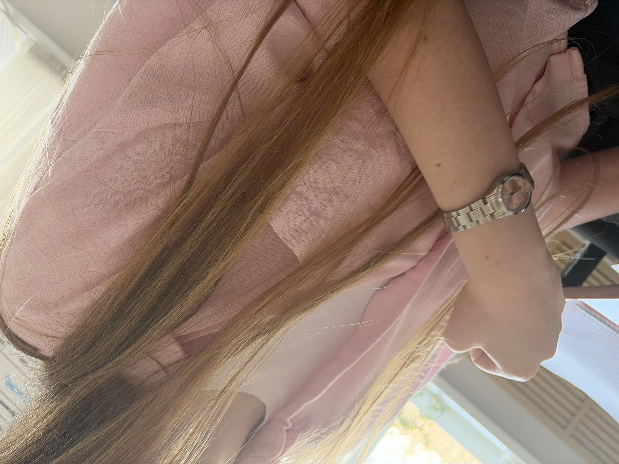 The image is a close-up, slightly angled view of a person wearing a pink shirt or jacket. The person has long, light brown hair that partially obscures the view. On their left wrist, they are wearing a silver wristwatch. The background is blurry, but it appears to show a window with some greenery visible outside. The location, Chaikovsky, Russia, cannot be determined from this image alone.