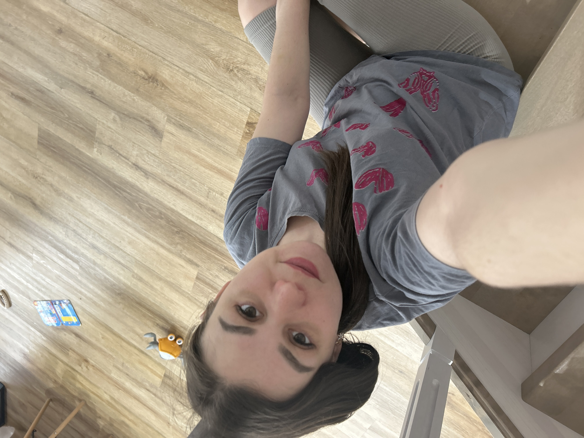 The image, rotated, captures a selfie of a woman lying or sitting on a light wood-patterned floor. Her head is positioned at the bottom right of the frame, with her gaze directed towards the camera, showing a neutral to slightly pleasant expression. She has dark hair and is dressed in a grey t-shirt featuring repetitive pink, stylized designs resembling horse or zebra heads. She also wears light grey ribbed leggings or pants, with her legs bent.
The setting is indoors, characterized by clean, light-colored wooden floorboards laid in a diagonal pattern across most of the frame. Several items are visible on the floor: a small orange and white plush toy shaped like a crab with prominent eyes, located near the woman's head. Further to the left, a small rectangular object, possibly a children's book or game card, displays colorful illustrations in blue and yellow. A light-colored ring or circular object is also present. On the far right, a section of a light-toned banister or stair railing suggests a residential interior, possibly near a staircase. The room appears brightly lit. No distinct text is clearly legible on any of the objects.