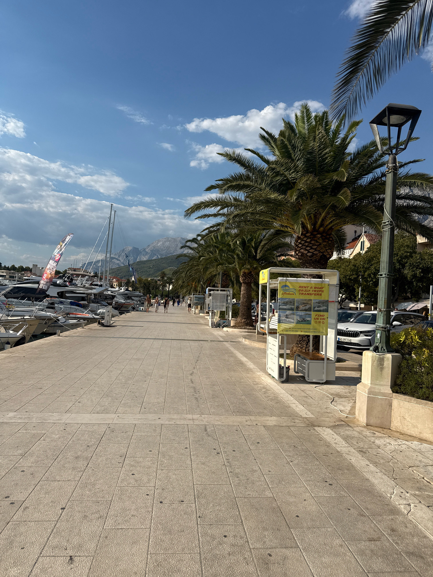 The image depicts a sunny day in the city of Makarska, Croatia, specifically along its waterfront promenade. The foreground features a paved walkway with a repeating pattern of rectangular stone tiles. To the left of the walkway, a marina is filled with various boats, including sailboats and smaller motor yachts. A tall, narrow flag with colorful graphics is planted on a boat. Palm trees line the right side of the walkway, casting dappled shadows on the ground. In the mid-ground, several people are walking along the promenade. A white kiosk with a yellow sign advertising boat rentals and transfers stands prominently on the right side of the walkway. Behind the kiosk, a white car is parked. In the background, buildings and lush green trees are visible, with a range of mountains forming a scenic backdrop under a bright blue sky dotted with scattered white clouds. A street lamp with a dark shade is positioned next to the kiosk. The overall scene conveys a relaxed atmosphere characteristic of a coastal resort town.