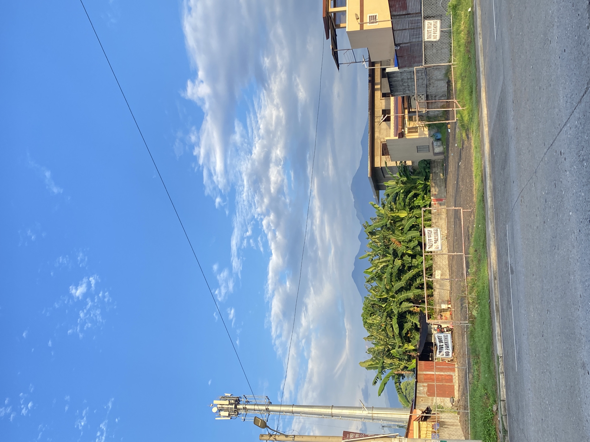 This is a medium shot of a scenic view in Davao City, Philippines, on a clear day with scattered clouds. The foreground shows a paved road and a grassy area. In the mid-ground, there is a fenced-off lot with a "DRIVE WAY NO PARKING" sign visible on the fence, and a tall cell tower stands to the left. Behind the lot, there are banana trees and buildings, including a yellow building with a balcony. In the background, a mountain range is visible under the bright blue sky.