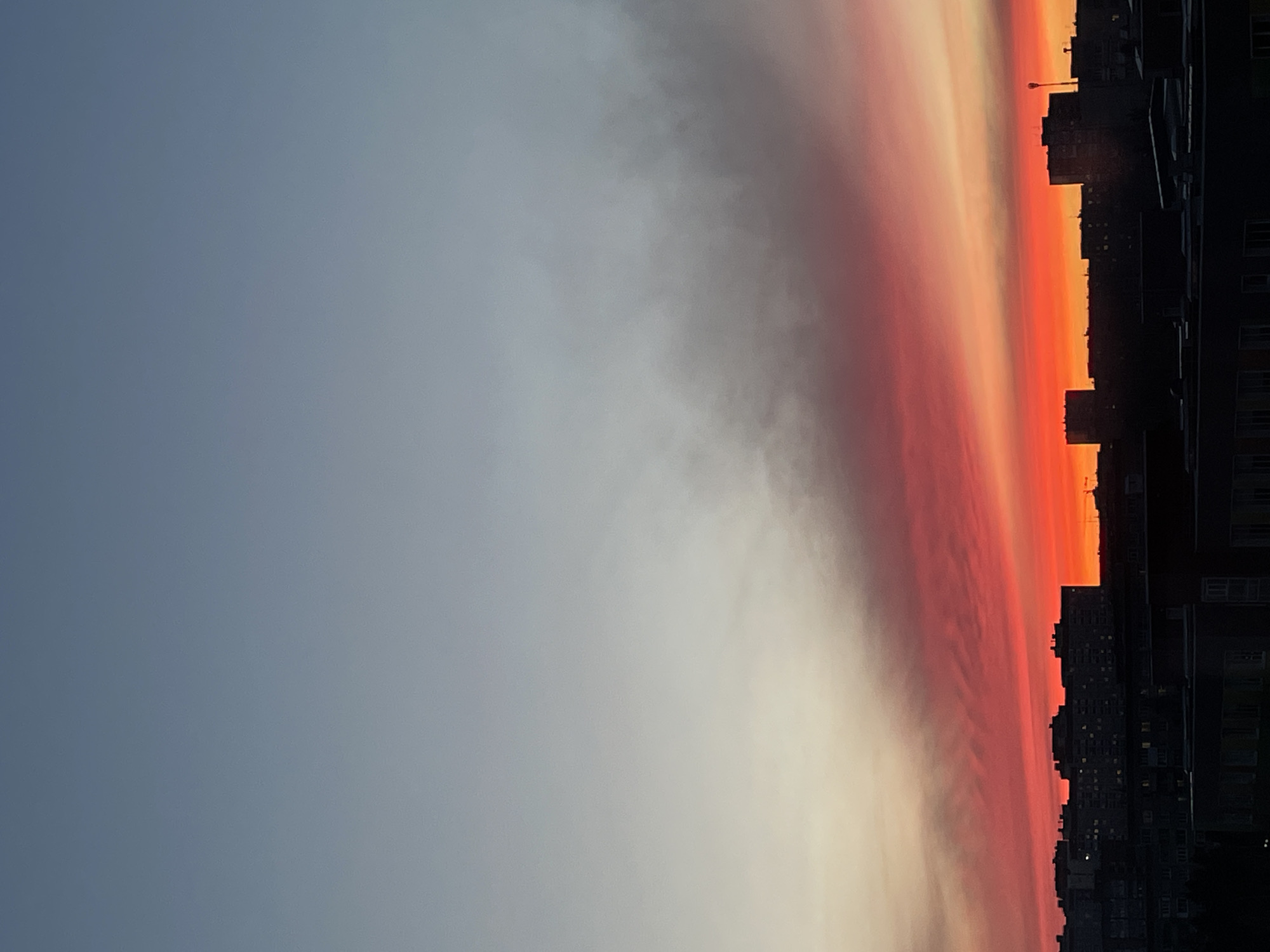 The image shows a sunset over the city of Izhevsk, Russia. The sky transitions from a light gray-blue to a vibrant orange and red near the horizon. The silhouettes of buildings are visible against the colorful sky.