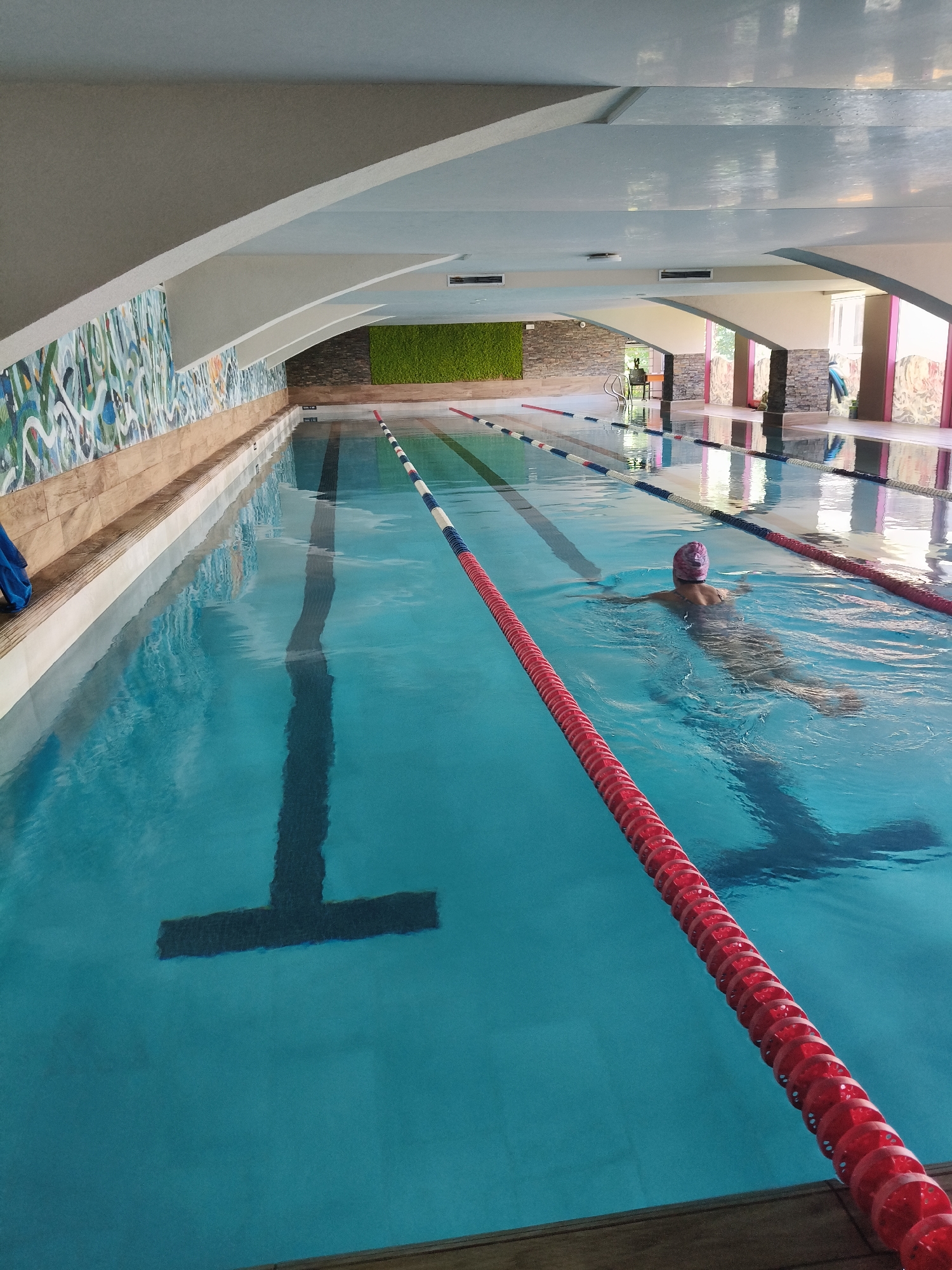 The image depicts an indoor swimming pool with several lanes marked by ropes. A person wearing a pink swimming cap is swimming in one of the lanes. The pool area has decorative walls with colorful abstract patterns and textured stone accents. The ceiling is a light blue color with arched structures. The lighting appears to be artificial, and there are reflections on the water's surface. This scene is located at the indoor swimming pool of the "Kolhozen Pazar" complex in Plovdiv, Bulgaria.