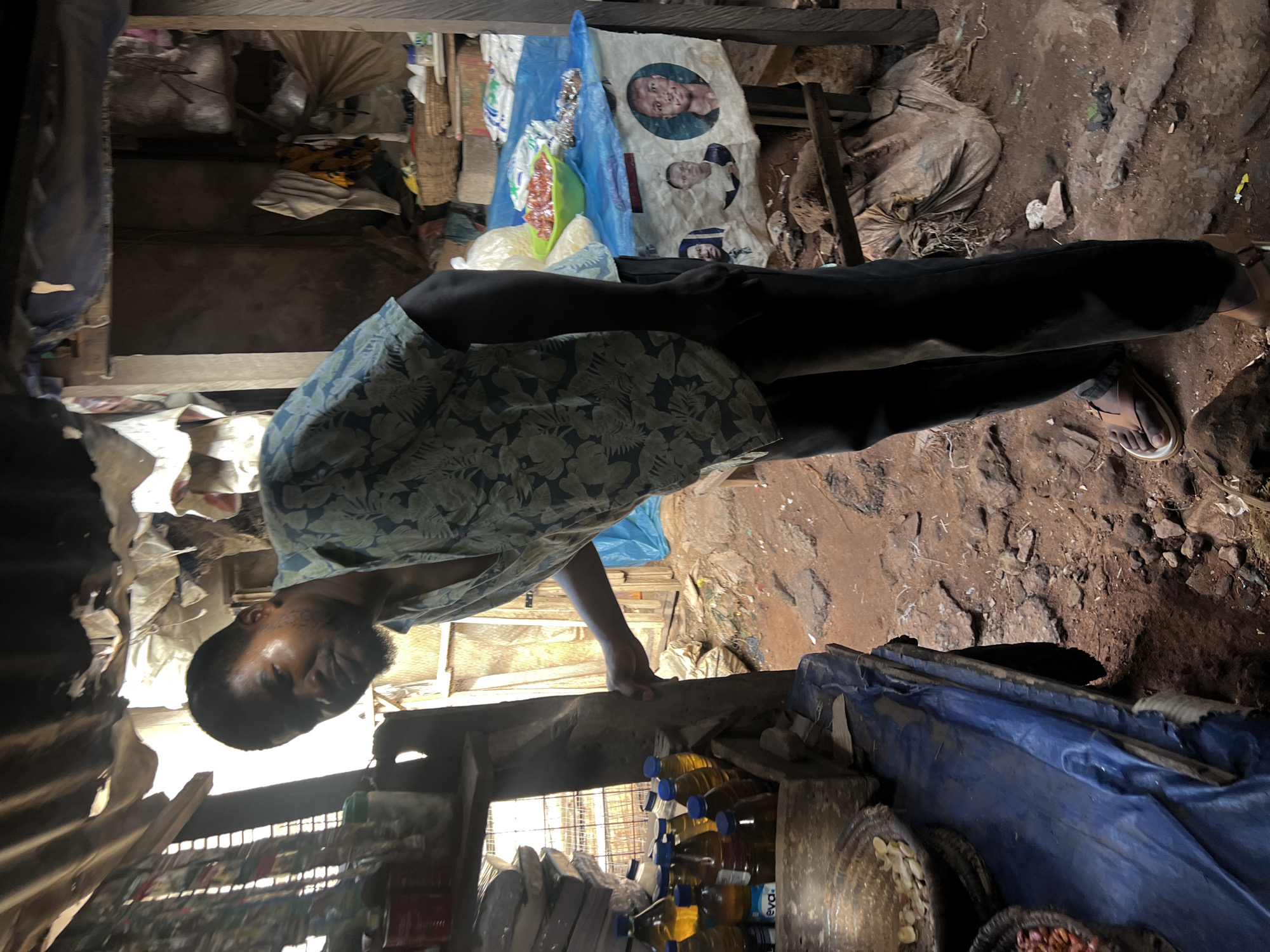 A man in a floral patterned shirt and dark trousers stands in a cluttered, dimly lit space, possibly a small shop or storage area. He appears to be inspecting the surroundings, leaning forward. The environment is filled with various goods, including what seems to be cooking oil in plastic bottles, sacks of produce, and blue tarpaulins. The ground is dirt. The precise location in the city of Unknown, Unknown cannot be determined from the image.