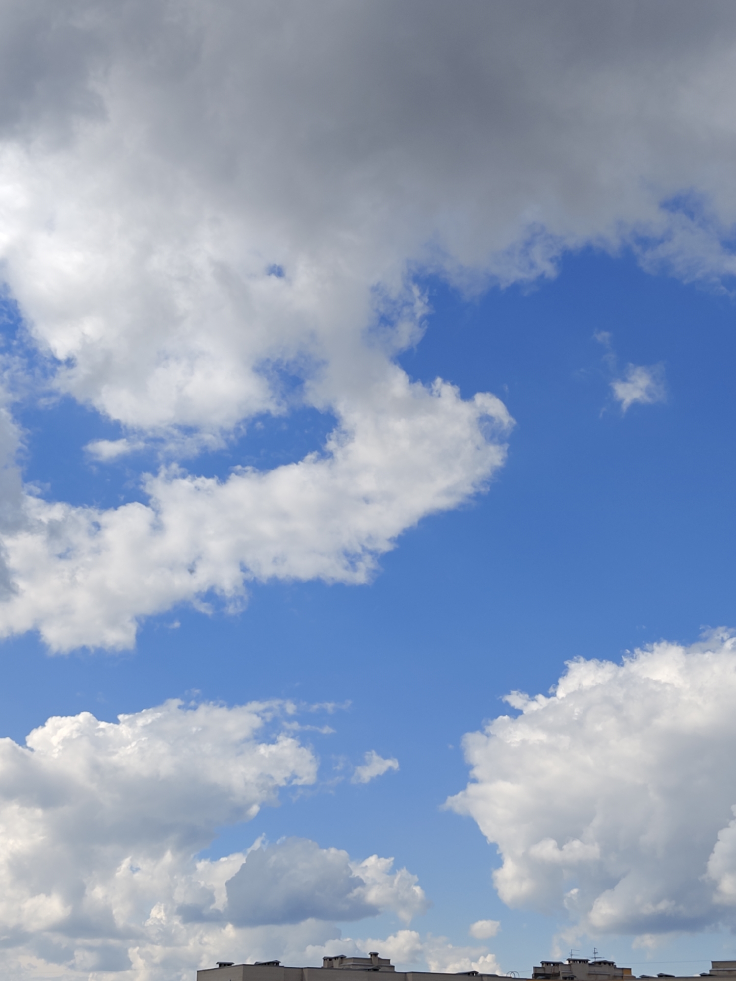 The image depicts a view of the sky from a city. The sky is bright blue with a mix of white and grey clouds. The clouds are fluffy and scattered, with some appearing more dense and dark. In the lower portion of the image, the rooftops of several buildings are visible, indicating an urban setting. The architecture suggests it could be part of Krasnoobsk, Russia, given the prevalence of apartment blocks in the region. There are no people or specific actions visible in the frame, focusing entirely on the atmospheric conditions and the city's skyline.