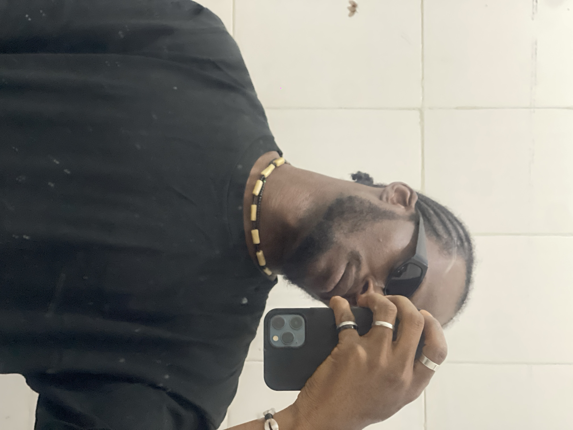 A dark-skinned male subject is depicted in a mirror selfie, taken indoors in Jos, Nigeria. He has dark hair styled in short dreadlocks or cornrows, a trimmed beard, and mustache. He wears dark, angular sunglasses that obscure his eyes. He is wearing a black short-sleeved t-shirt, a two-strand beaded necklace featuring light-colored cylindrical beads and small dark beads, and multiple rings on his left hand. These include two wider silver-colored bands on his index and ring fingers, and a thinner black band with a silver stripe on his pinky finger. A cowrie shell bracelet is faintly visible on his right wrist. The subject holds a black smartphone, featuring a triple-lens camera module, in his left hand to capture his reflection. The phone is encased in a plain black cover. The background consists of light-colored tiled walls with visible darker grout lines, creating a grid pattern. A small, reddish-brown insect-like detail and a tiny pink dot are present on the upper left wall section.