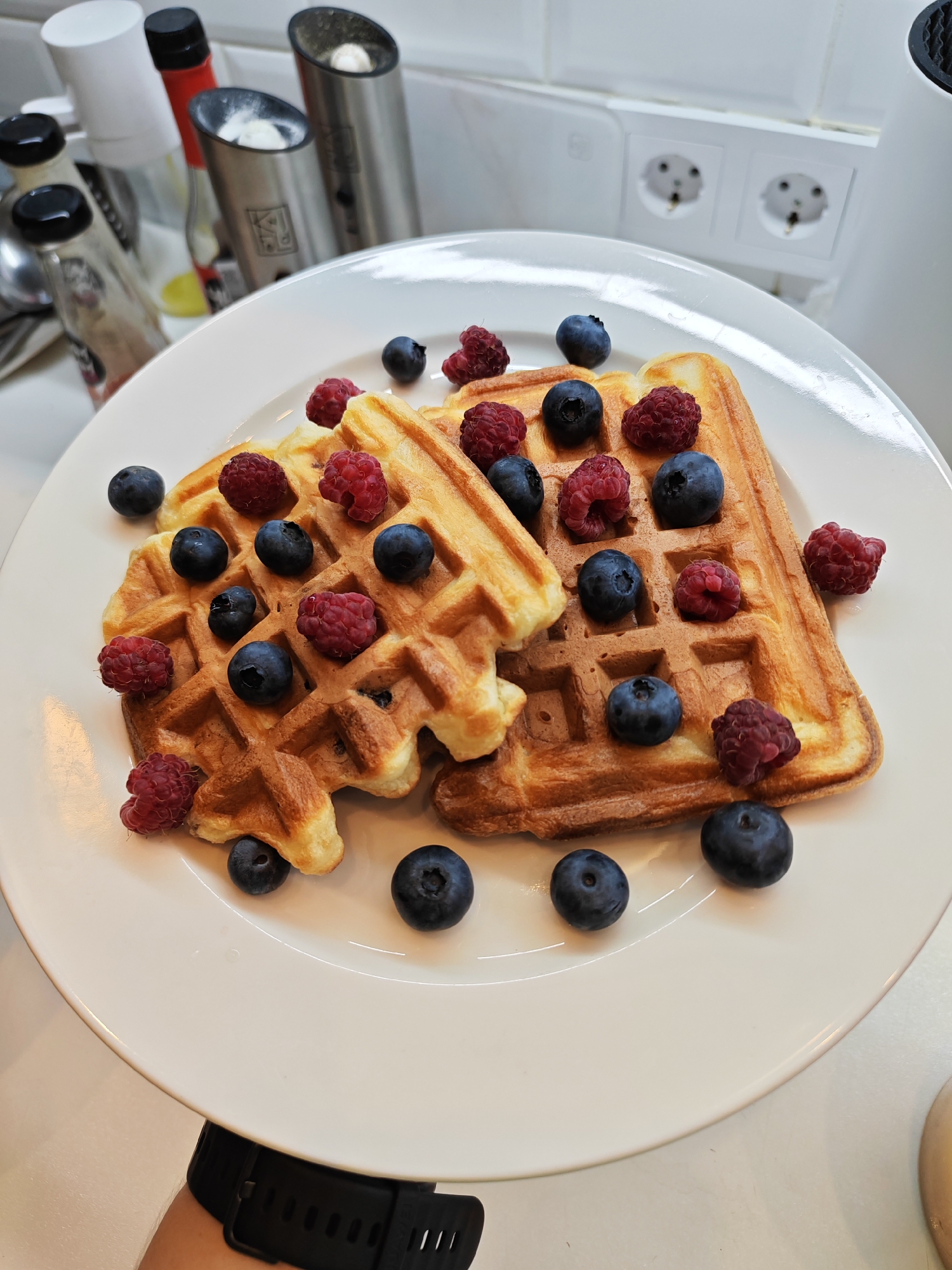 Two golden-brown waffles topped with fresh blueberries and raspberries sit on a white plate. The plate is positioned on a white countertop, with kitchen appliances and electrical outlets visible in the background. A person's arm, wearing a black smartwatch, is partially visible at the bottom of the image. The scene appears to be indoors, likely in a kitchen.