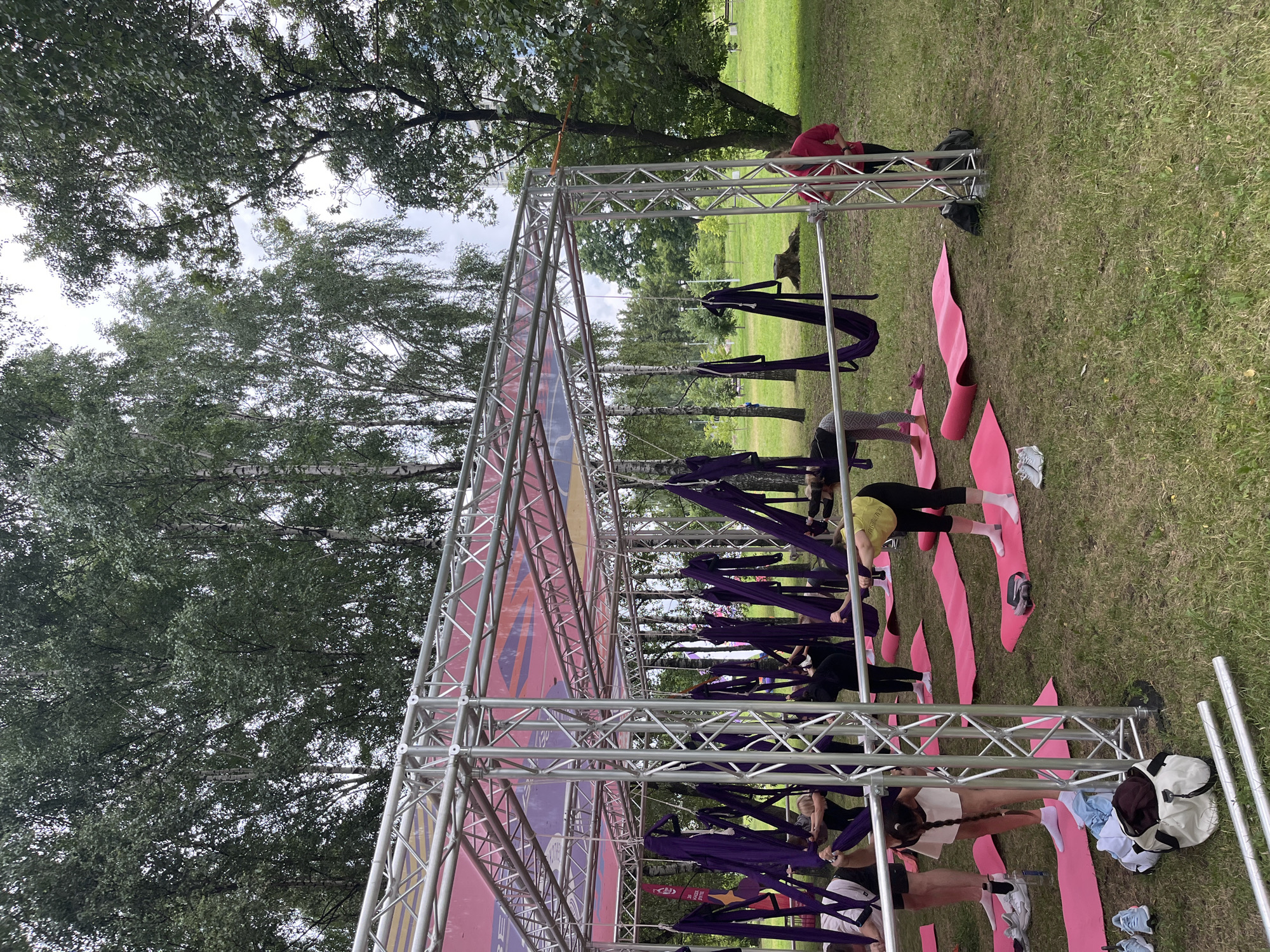 In an outdoor park setting, likely in the city of Minsk, Belarus, a group of individuals are participating in an aerial yoga class. The scene is set under a metal truss structure supporting a canopy with pink and purple banners. Several individuals, predominantly women, are engaged in yoga poses on pink mats spread across the grassy ground. They are using purple fabric slings, suspended from the truss, for support and various poses. The individuals are dressed in athletic wear. The environment is green and lush with mature trees in the background, suggesting a park or recreational area. The sky is overcast, indicating a cloudy day. Among the subjects, one person in a bright pink top stands to the right, observing the class, while others are actively performing yoga. Scattered on the grass are personal belongings such as a white duffel bag and athletic shoes.