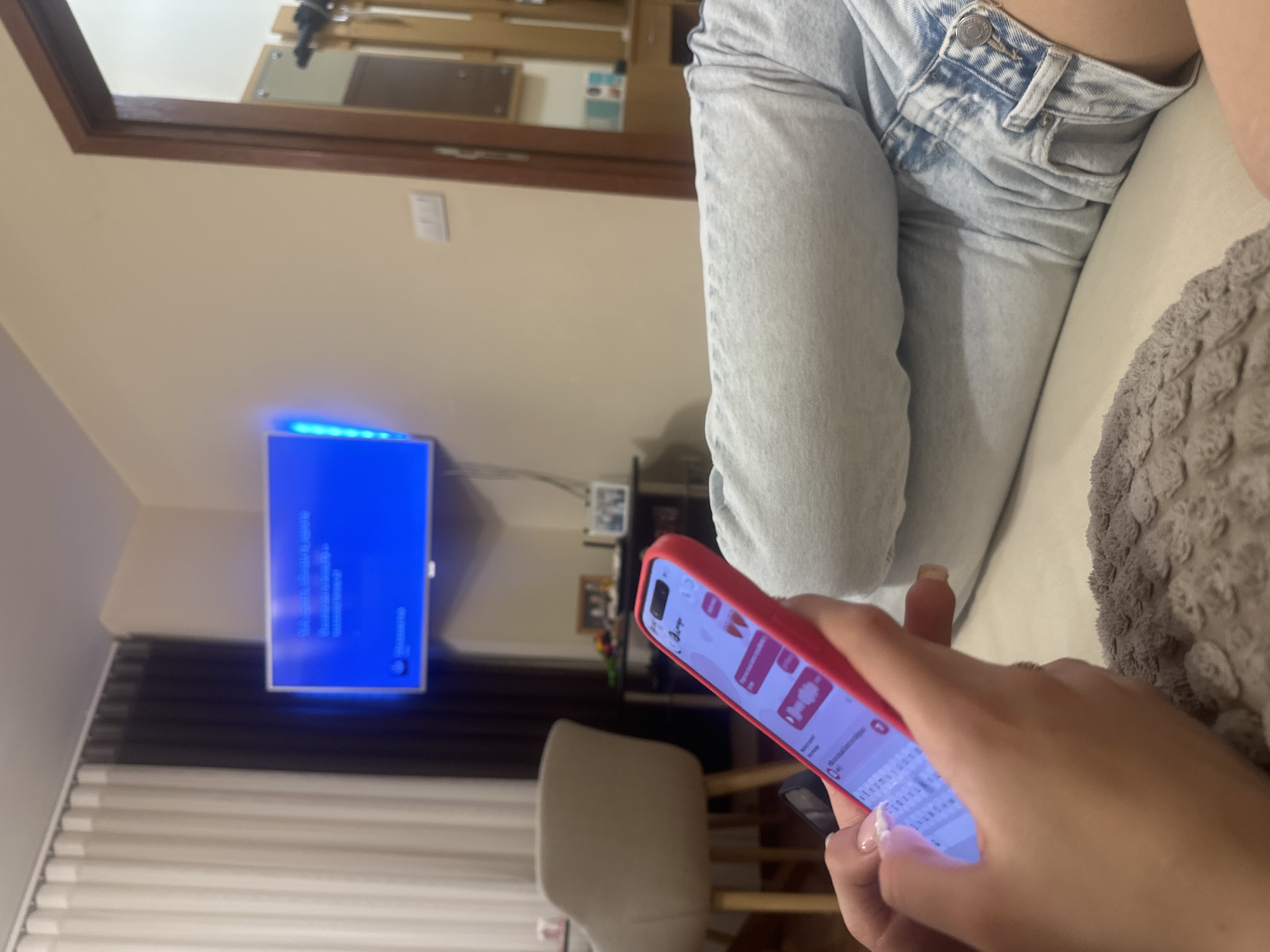 The image depicts a close-up view of a person's hands holding a pink-cased smartphone. The phone screen displays a messaging application with conversation bubbles and keyboard. The person is wearing light blue denim jeans. In the background, a television screen displays a bright blue interface. To the left, a chair with light-colored upholstery is visible, along with floor-to-ceiling curtains, one white and one dark gray. Further in the background, a doorway with a wooden frame leads to another room, and a mirror reflects some objects. The scene appears to be an indoor residential setting. The location is Shumen, Bulgaria.