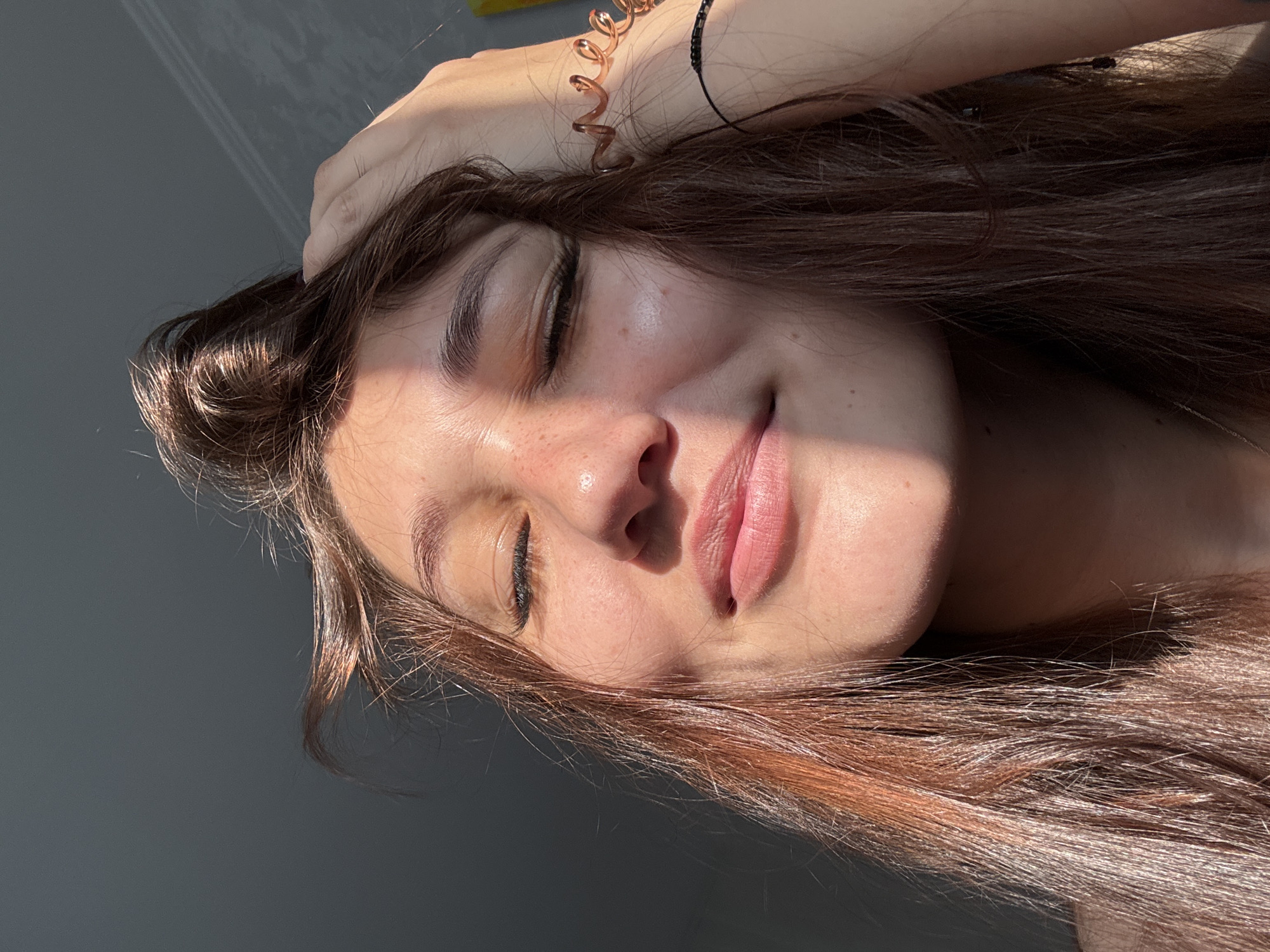 A close-up, low-angle outdoor portrait shows a young woman with her eyes closed and a slight smile. She has long, brown hair that is pulled up on one side with her left hand. Her face is partially illuminated by sunlight, casting a shadow across her nose and cheek. She has light makeup, including mascara and lipstick. There are a few freckles visible on her cheeks. She is wearing a rose gold spiral bracelet on her right wrist and a thin black beaded bracelet below it. The background is a plain, dark gray wall. There is no visible indication of the city of Novokuznetsk, Russia, or any specific location within it.