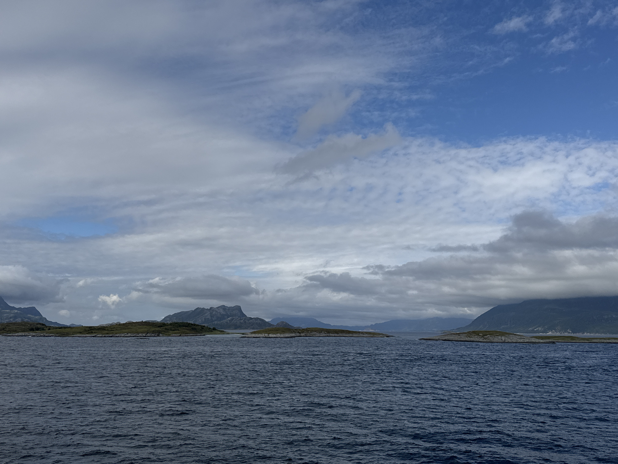 The image shows a wide body of dark blue water in the foreground, with small waves creating a textured surface. In the middle ground, several small, rocky islands with green vegetation dot the water. Behind the islands, a range of mountains can be seen, some with jagged peaks and others appearing more rounded and covered in vegetation. The sky above is a mix of blue sky and scattered clouds, some of which are dark and heavy, while others are wispy and white. The overall impression is of a rugged, natural landscape. The scene is located in Stokkvågen, Norway.