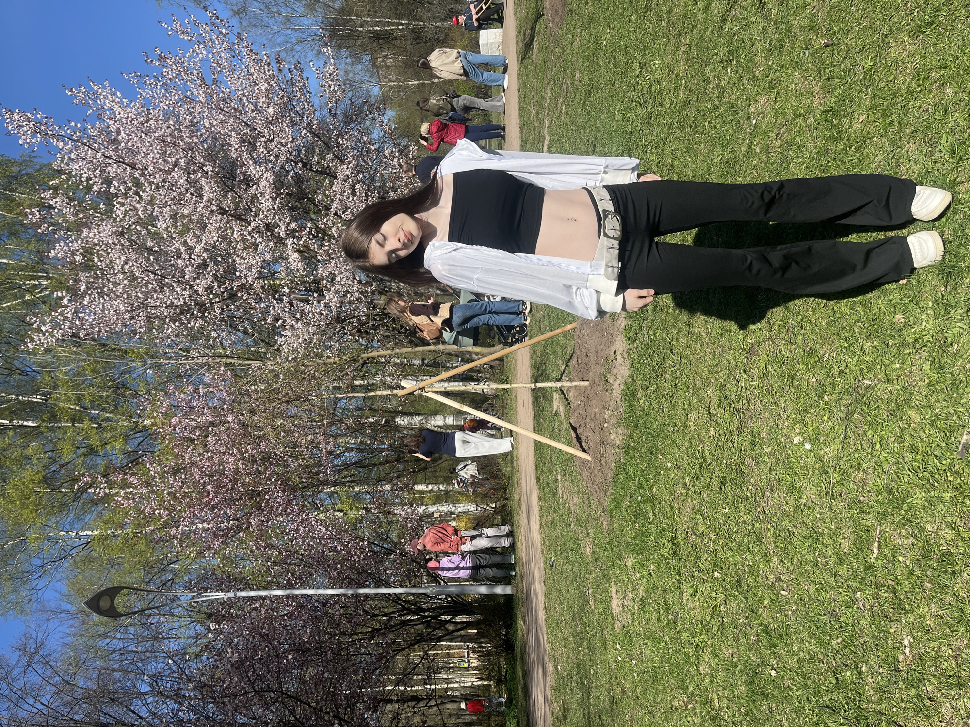 The image captures a young woman lying on a grassy area, possibly a park. She wears a black top, black pants, a white shirt, and a white belt. Behind her is a flowering tree with pink blossoms. In the background, other people are visible, suggesting the scene is in a public park. A lamppost is visible at the bottom left. There is also a wooden structure in the background.