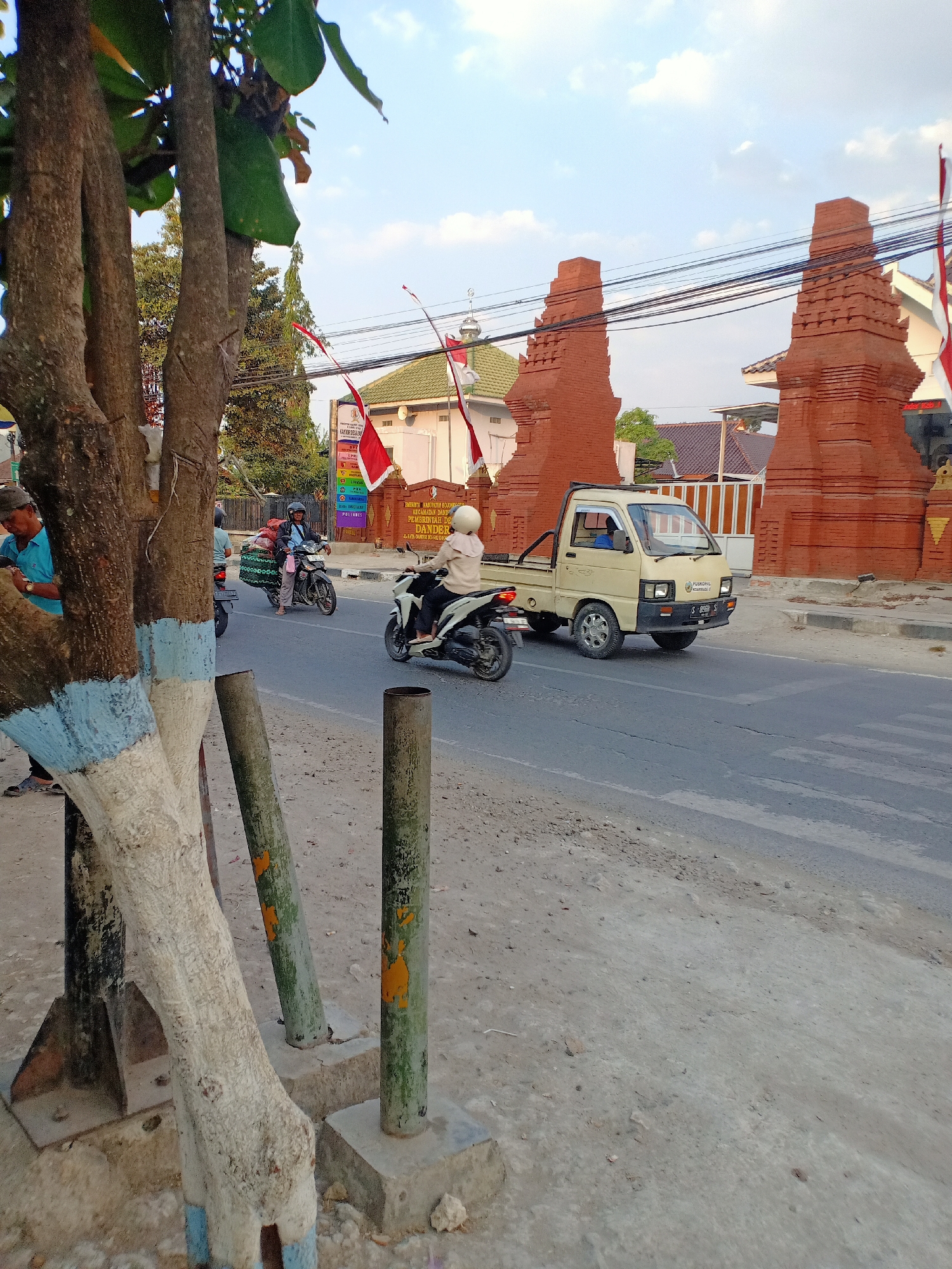 In Sumber Tlaseh, Indonesia, a street scene unfolds under a clear sky. The foreground features a textured tree trunk, partially painted with blue and white bands, and several weathered poles. A road curves through the mid-ground, occupied by a light-colored pickup truck and two motorcycles. The truck, driven by a person in blue, heads away from the viewer. A motorcyclist in a white helmet, with a tan jacket and dark pants, rides towards the viewer, while another motorcycle, carrying a load of goods, moves away from the viewer. The background is dominated by traditional red brick gateway structures. Behind them, a building with a green roof, possibly a mosque, is visible, adorned with red and white flags. Utility wires crisscross the sky above the gateway. Several people are visible, including one in a blue shirt and a cap standing near the tree, and the drivers of the vehicles. The overall environment appears to be a street in a populated area, likely a town or city.