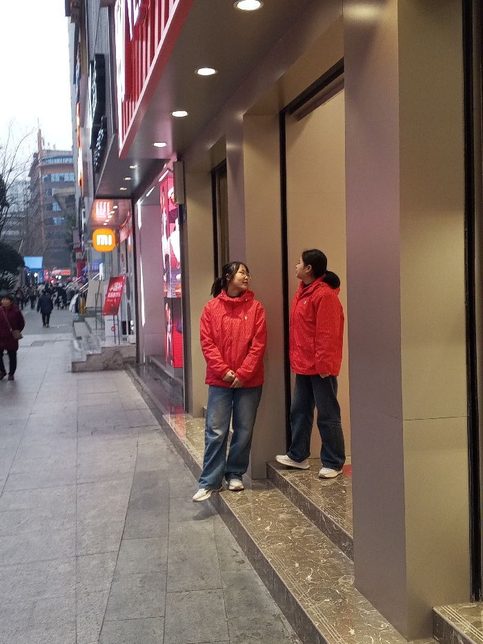 The image depicts an urban street scene in Jiangjin, China, under an overcast sky. In the foreground, two East Asian females, appearing to be young adults, are positioned on the right side of the frame near a commercial building entrance. Both individuals wear identical red hooded jackets and blue jeans.
The female on the left stands on the paved sidewalk, wearing glasses, with her dark hair tied back, and her hands clasped in front of her. She is looking towards the right, at the second female. The female on the right stands on the top of two shallow, brown-patterned tiled steps leading into the building. She also has dark hair tied back and is looking towards the left, at the first female. Their gazes suggest an interaction or conversation.
The building facade behind them features a red-colored horizontal structure with multiple recessed lights. To the left of the main subjects, a prominent orange "Mi" (Xiaomi) logo sign is visible on the building, indicating a retail establishment. Red promotional banners are also present near the storefront. The sidewalk, constructed from grey paving slabs, extends into the mid-ground on the left, where several other blurred pedestrians are visible walking. Multi-story urban buildings line the street further in the background.