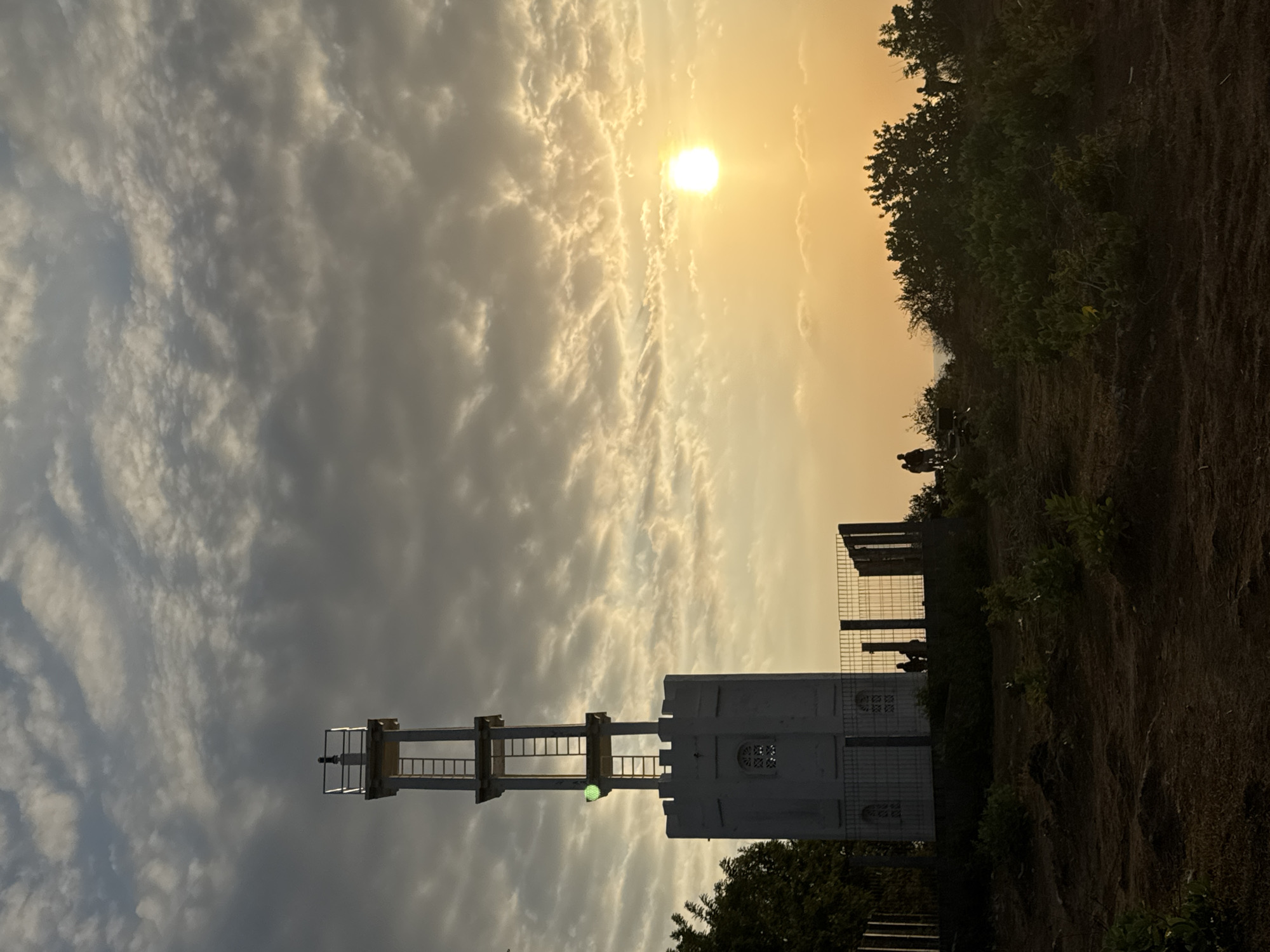 The image depicts a lighthouse structure on a hilltop overlooking a sunset in Kuta Selatan, Indonesia. The lighthouse is white with a square base and a tower that extends upwards, featuring a ladder and railings. A green light is visible on the side of the tower. In the foreground, there are some bushes and uneven terrain. In the midground, several silhouetted figures of people are gathered, some near the lighthouse and others further away, appearing to observe the sunset. The sky is filled with dramatic clouds, illuminated by the bright, setting sun which is positioned to the right of the frame, casting a warm, golden glow over the scene. The environment appears to be a coastal or elevated area with vegetation.