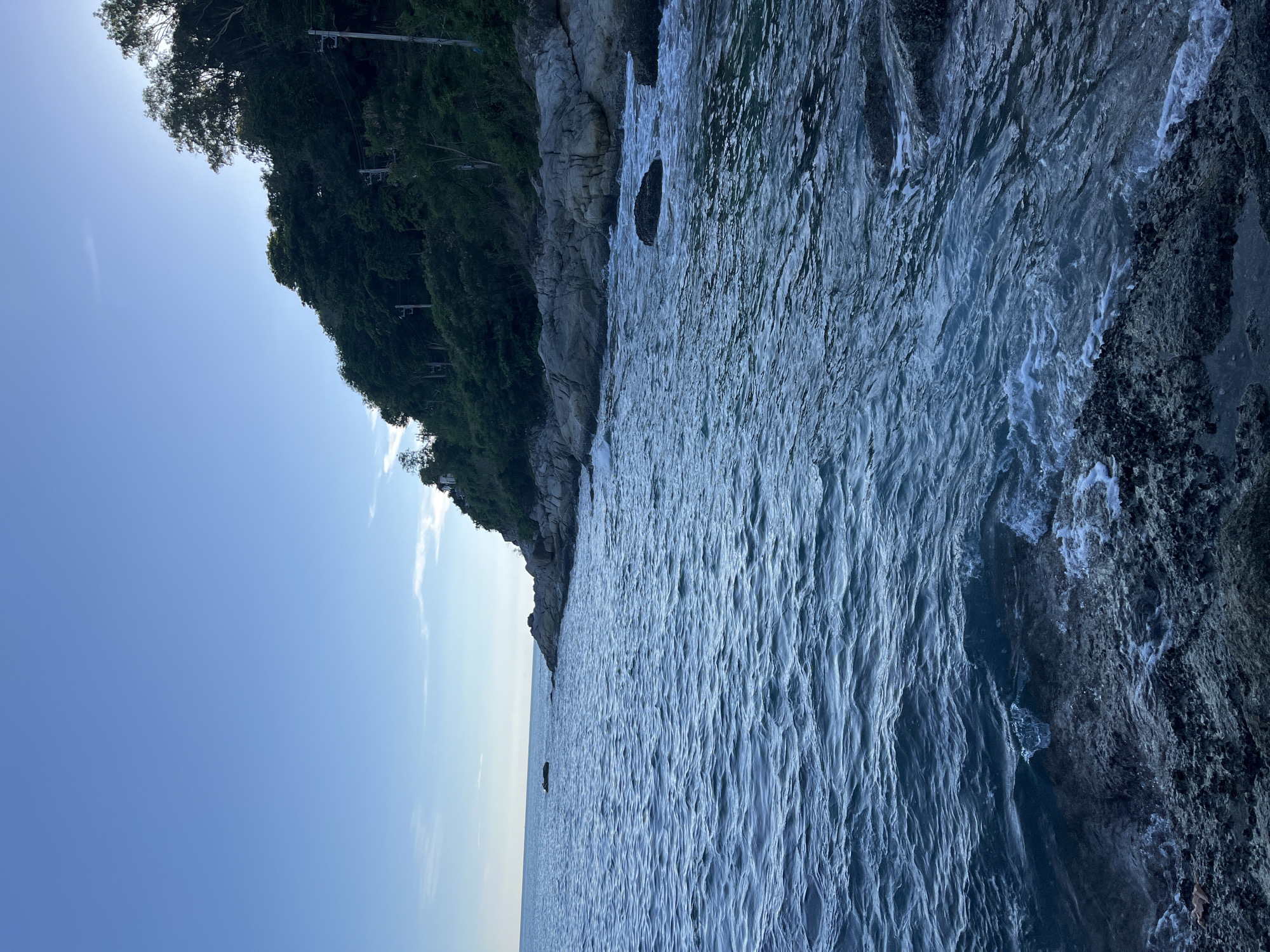 The image depicts a rugged coastline with choppy, dark blue ocean waves crashing against a rocky shore. A densely wooded hill rises steeply from the coast, with a clear blue sky above. In the distance, a small, dark object appears to be floating on the water. Electrical poles are visible amongst the trees on the hillside. The overall scene suggests a natural, undeveloped coastal area. The location is specified as Rawai, Thailand.