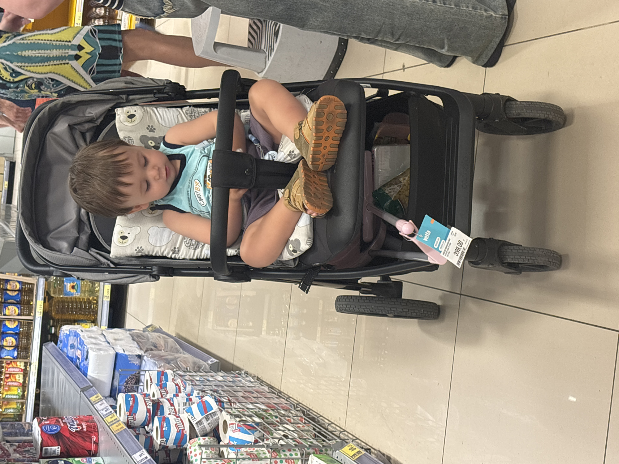 A young child is seated in a stroller in a supermarket aisle. The child is wearing a blue tank top and grey shorts, with brown shoes. The stroller has a patterned cushion. To the left of the stroller are shelves stocked with toilet paper and other household items. In the background, another person is partially visible wearing a patterned dress and jeans. The floor is tiled. There is a price tag visible on the stroller. The scene is consistent with a shopping experience in a supermarket in Sochi, Russia.
