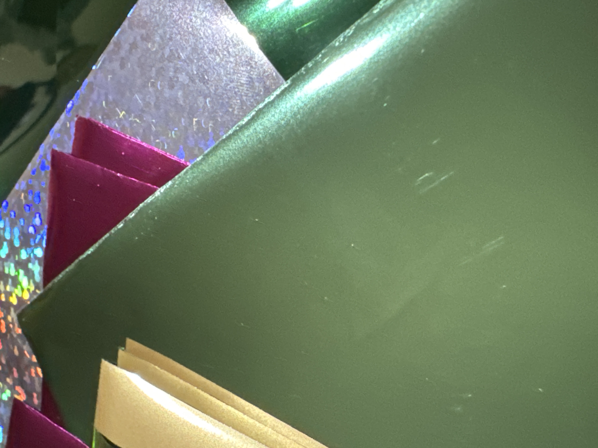 The image is a close-up, angled shot of several pieces of what appears to be decorative wrapping paper. The dominant color is a deep, glossy green, which takes up the majority of the frame and shows a slight sheen. To the upper left, there are stacked strips of gold-colored paper, also with a subtle sheen. Further up and to the right, layered beneath the gold, are pieces of bright pink and holographic paper that reflects a rainbow of colors. A sliver of dark green, glossy material is visible on the far right edge of the frame. The surface of the green paper shows a few faint scratches and smudges. There are no people, animals, or discernible city landmarks present in the image. It is not possible to determine the location in Waterford, Ireland, as the image contains no identifying features.