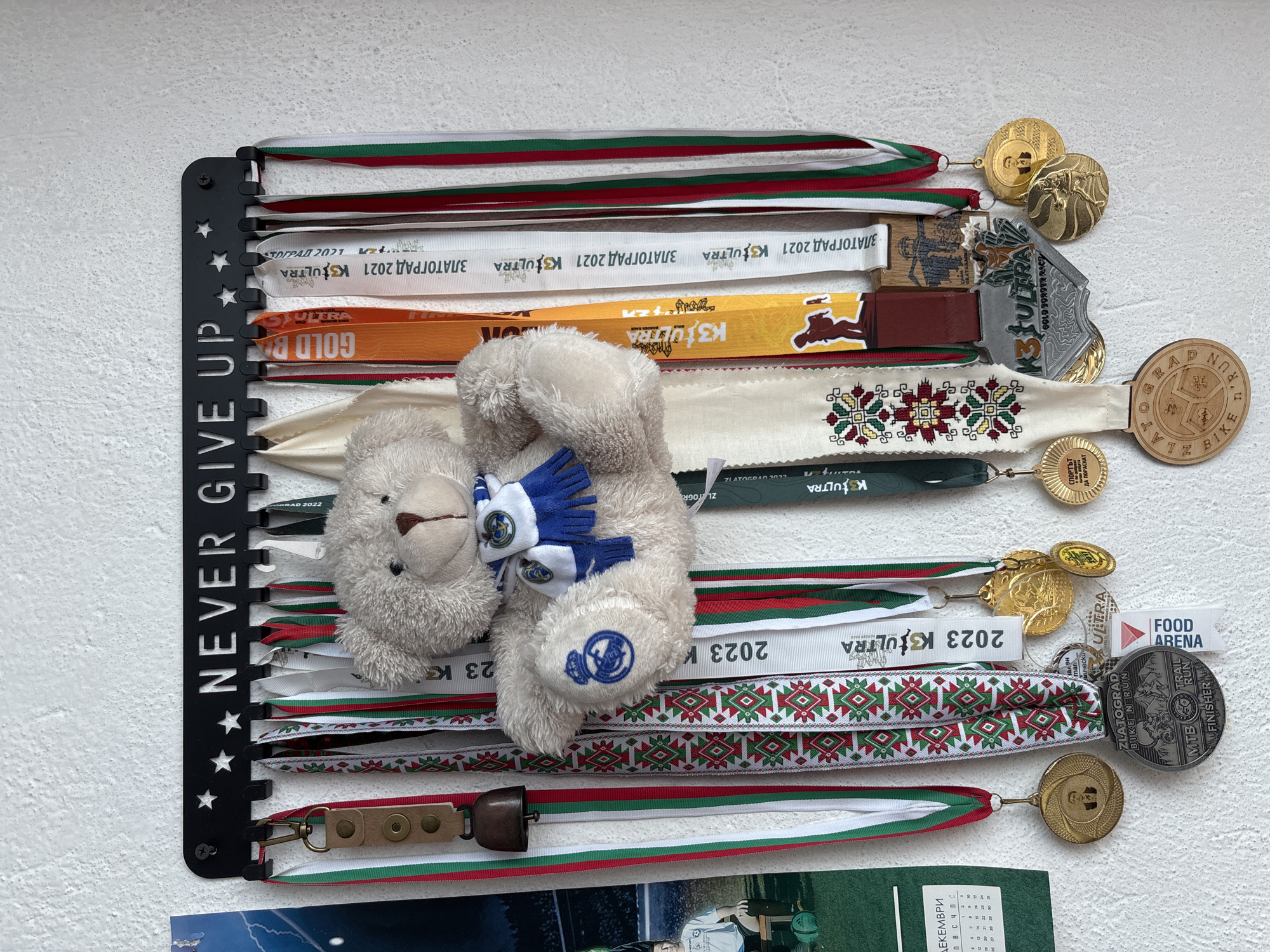 The image displays a medal hanger with the phrase "NEVER GIVE UP" and star cutouts along its side, mounted on a textured white wall. A plush teddy bear with a blue and white scarf is positioned in front of the medals.
The medals and ribbons are displayed on the hanger. There are multiple ribbons in the colors of the Bulgarian flag (white, green, and red), as well as ribbons with event names like "ZLATOGRAD 2021 K3ULTRA" and "2023 K3ULTRA". Some medals are attached to the ribbons, including gold, silver, and wooden medals with engravings related to Zlatograd bike and run events. One medal is specifically marked "FOOD ARENA" and "ZLATOGRAD BIKE N RUN".
A calendar displaying the month of "ДЕКЕМВРИ" (December) is partially visible at the bottom of the frame.