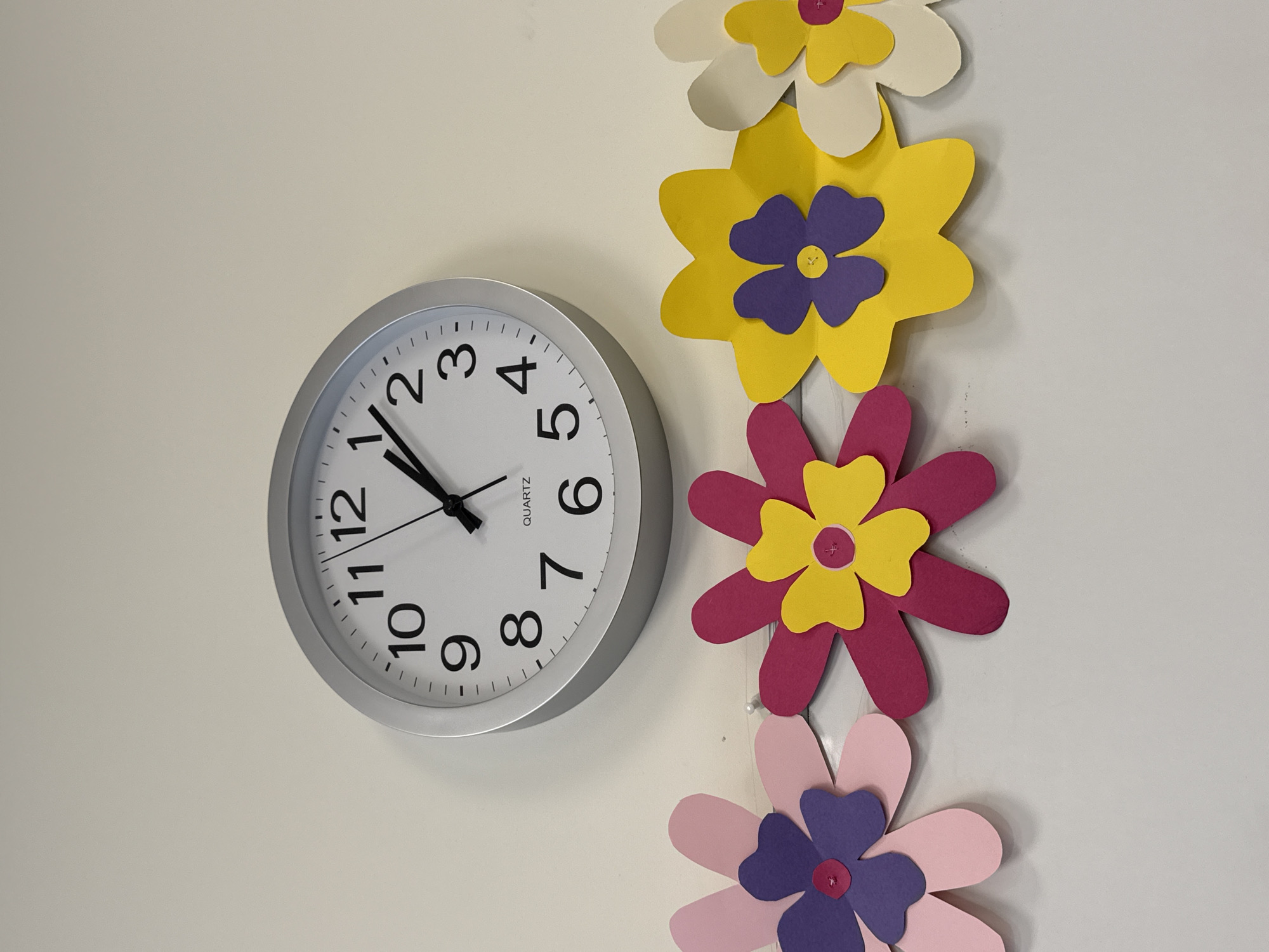 A clock and decorative paper flowers are attached to a plain white wall. The clock is round with a silver frame, white face, and black numerals. The hands of the clock indicate a time of approximately 10:52. The paper flowers are arranged vertically next to the clock. The top flower is white with a yellow center, followed by a yellow flower with a purple center, a pink flower with a yellow center, and a light pink flower with a purple center. The location is Unknown, Unknown.