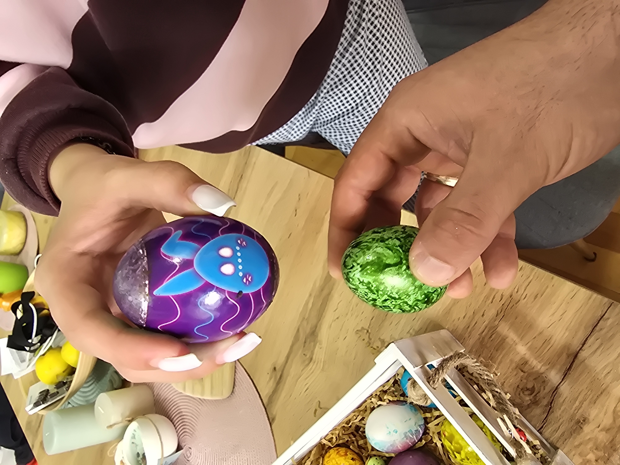 This close-up, overhead shot captures two individuals' hands interacting with decorated Easter eggs on a light wooden table in Burgas, Bulgaria. The person on the left, wearing a long-sleeved top with broad pink and maroon stripes, holds a vibrant purple Easter egg between fingers with white-tipped, manicured nails. This egg features a playful blue rabbit design, accented with wavy lines and a glittery silver section. Adjacent to it, the hand of another person, wearing a checkered shirt and dark pants, holds a smaller, intricately patterned green Easter egg. A gold-colored ring is visible on this person's right ring finger.
The warm, light wooden table, with its visible grain, serves as the backdrop for this Easter-themed activity. In the lower right corner, a white wooden crate contains several other decorative eggs, nestled in straw-like packing material. These include a marbled light blue and white egg and a speckled yellow-orange egg, suggesting a variety of designs. On the left side of the frame, out-of-focus background elements include a small wooden bowl filled with yellow lemon-shaped items and other small trinkets, along with several light green and cream-colored pillar candles. The scene conveys a sense of festive preparation or appreciation of handcrafted items, likely related to an Easter celebration. No visible text is present in the image.
