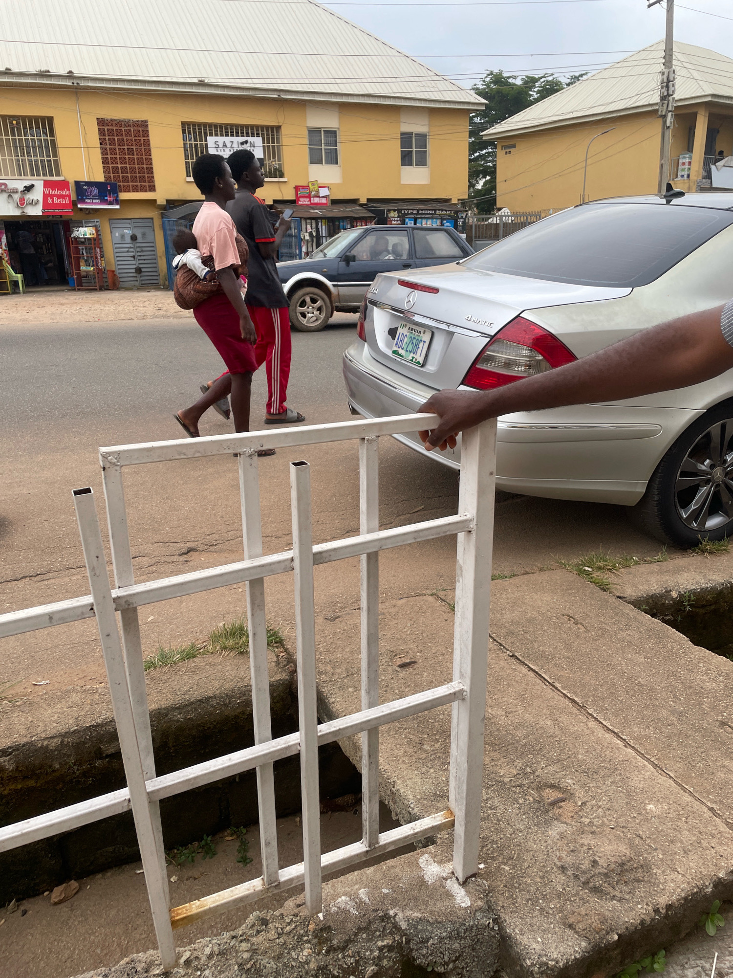 In Jos, Nigeria, two individuals walk along a paved street. A woman is seen from the side, carrying a baby on her back, wrapped in a brown fabric sling. She wears a pink t-shirt and a dark red skirt. Beside her, a person in a black t-shirt and red track pants walks, looking forward. A silver Mercedes-Benz sedan is parked on the right side of the street, partially obscuring the view of the sidewalk and a white metal railing. The railing appears to be part of a low barrier or structure along the edge of the sidewalk, which is made of concrete and shows signs of wear. In the background, yellow buildings with white roofs line the street, one of which has signs for businesses. The sky is overcast. A hand with dark skin is visible in the foreground, resting on the white railing.