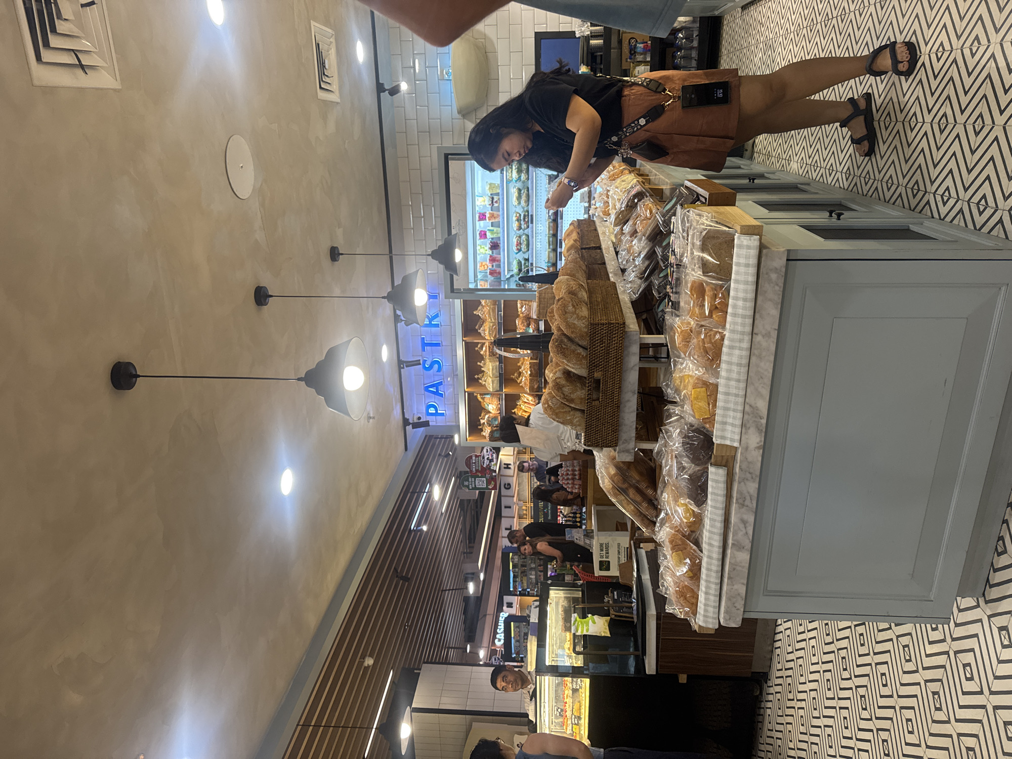 A bustling indoor pastry shop in Kuta Selatan, Indonesia, features a wide display of baked goods. The shop's interior has a modern, clean aesthetic with a light-colored concrete ceiling and white tiled walls. Hanging pendant lights illuminate the space, casting a warm glow on the products. A prominent "PASTRY" sign in neon blue letters is visible above a refrigerated display case. Several people are browsing the offerings: a woman in a black top and orange skirt is looking at her phone while standing near a display of packaged pastries. In the background, other customers are visible, including a cashier attending to a customer at a counter. The floor is covered in a striking black and white geometric tile pattern. Various breads, pastries, and pre-packaged food items are arranged on shelves and in baskets, inviting customers to make their selections.