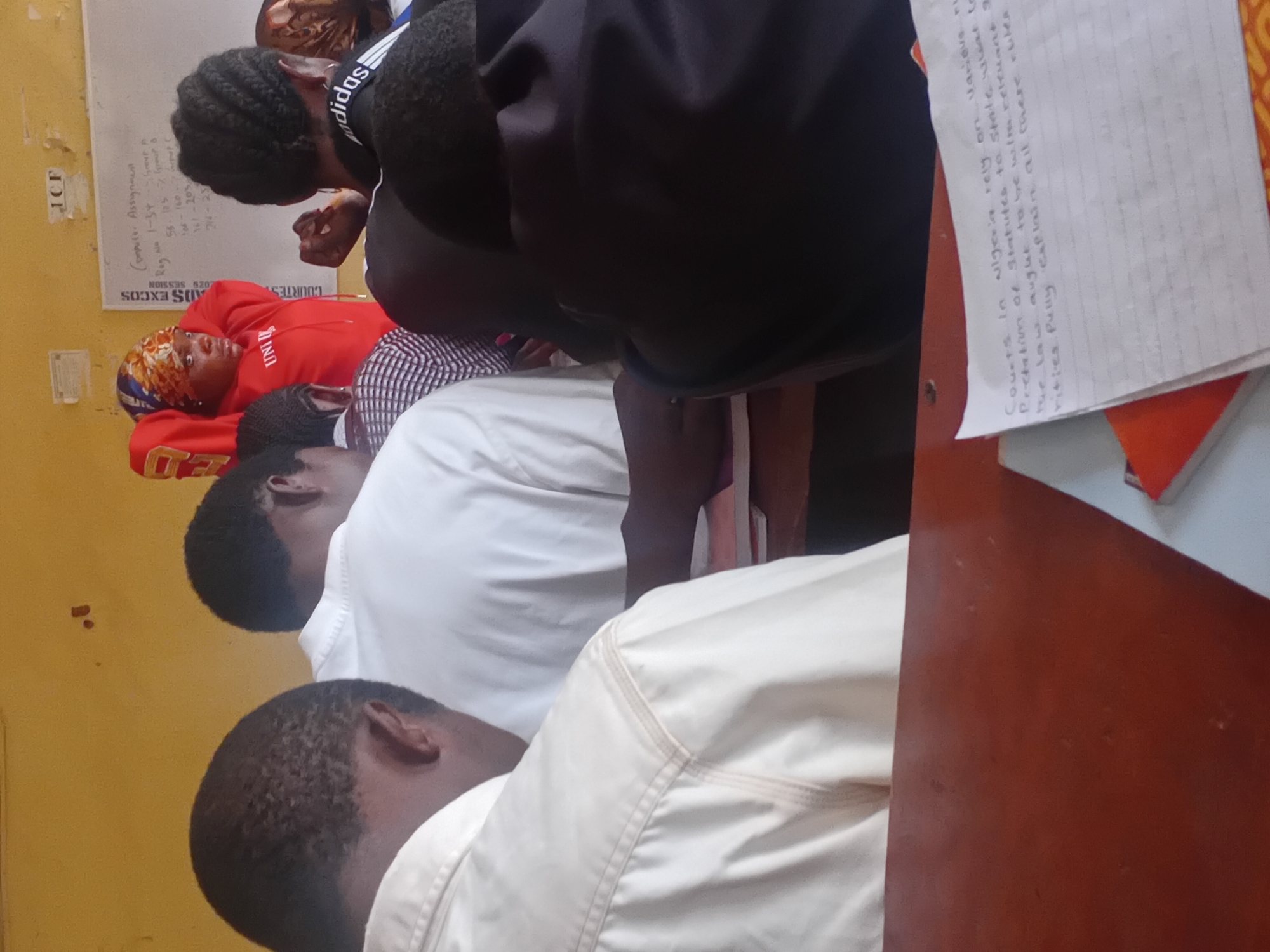 The image depicts an indoor scene, likely a classroom, in Ogbogbo, Nigeria. In the foreground, a wooden desk is visible. On the desk, a sheet of lined paper with handwritten text is placed on top of a stack of books. The text on the paper reads: "Courts in Nigeria rely on various r\nProclamation of Statutes to State rele\nThe law aught to be helm relevant t\nfitles fully explain all there rules".
Several individuals are present in the background, mostly seen from the back or side. One person, wearing a red hoodie and a patterned headscarf, stands near a whiteboard. The whiteboard has handwritten text and drawings, including what appears to be a list of assignments and student registration numbers. Other individuals, predominantly young men, are seated or standing, appearing to be students. Their attire suggests a casual or academic setting. The overall environment appears to be a modest educational institution, with yellow walls and a whiteboard as central features.