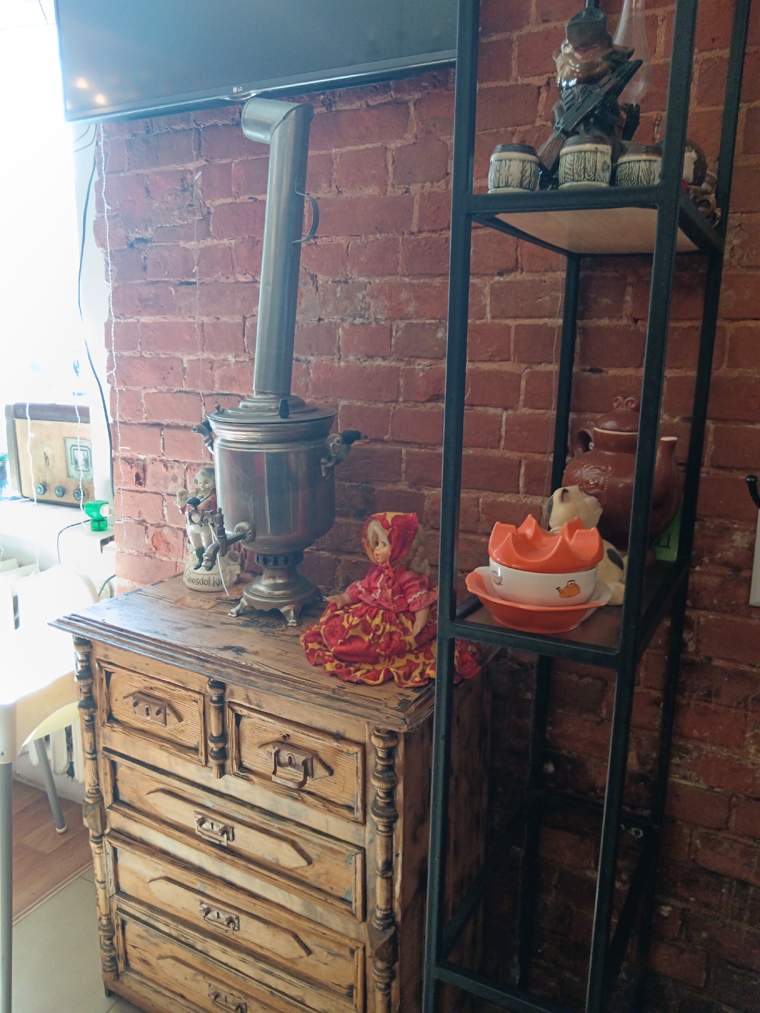An indoor scene depicts a vintage wooden dresser with a samovar, two small figurines, and a doll on top. To the right, a black metal shelf unit displays ceramic decorative items. The backdrop is a red brick wall, and a television is mounted above the dresser. The scene appears to be within a room in Gorokhovets, Russia, suggesting a domestic or shop environment with a collection of antique and decorative objects.