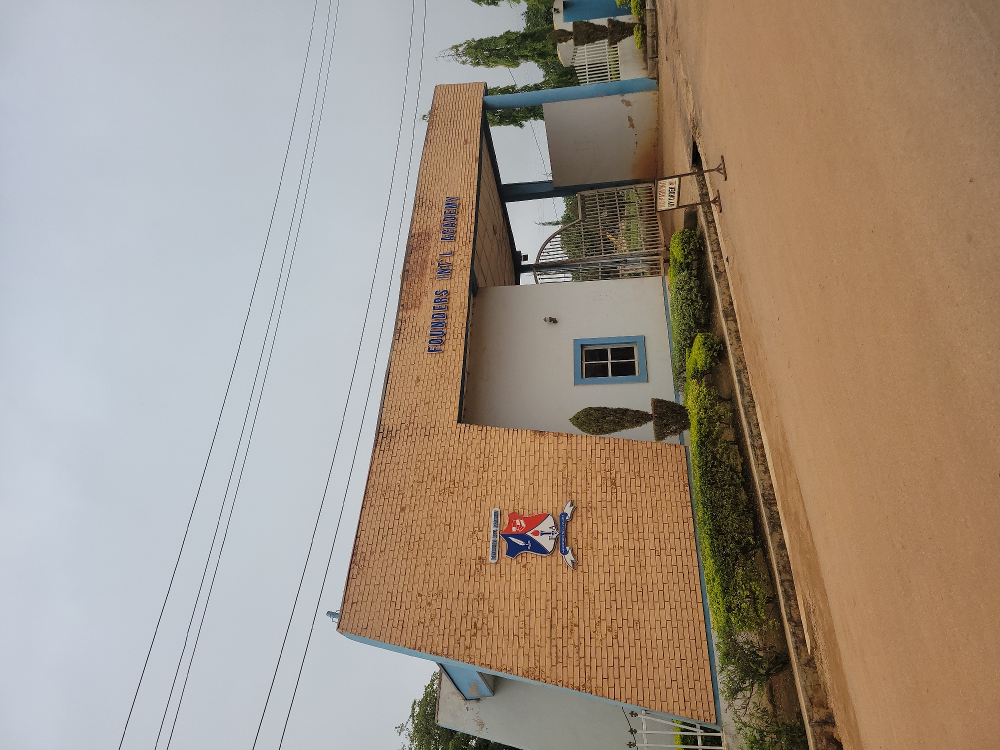 The image shows the entrance of "Founders Int'l Academy" in Barakin Akawo, Nigeria. The academy's name is prominently displayed in blue lettering above the entrance archway. The building features a distinctive architectural style with a beige brick facade that angles upwards to the left. A school crest, featuring a quill, a book, and a torch, is mounted on the brick section.
To the right of the brick section is a white, modern-looking structure that forms the main entrance. A large metal gate, partly open, leads into the school grounds, revealing a glimpse of trees and greenery within. A small, framed "NO PARKING BY ORDER" sign stands on the right side of the paved entrance area.
The academy is flanked by well-maintained green hedges and manicured shrubs, suggesting a cared-for environment. Tall, slender trees are visible in the background, and the sky is overcast, indicating a cloudy day. The foreground is a wide, light brown paved road. The overall scene depicts the entrance to an educational institution.