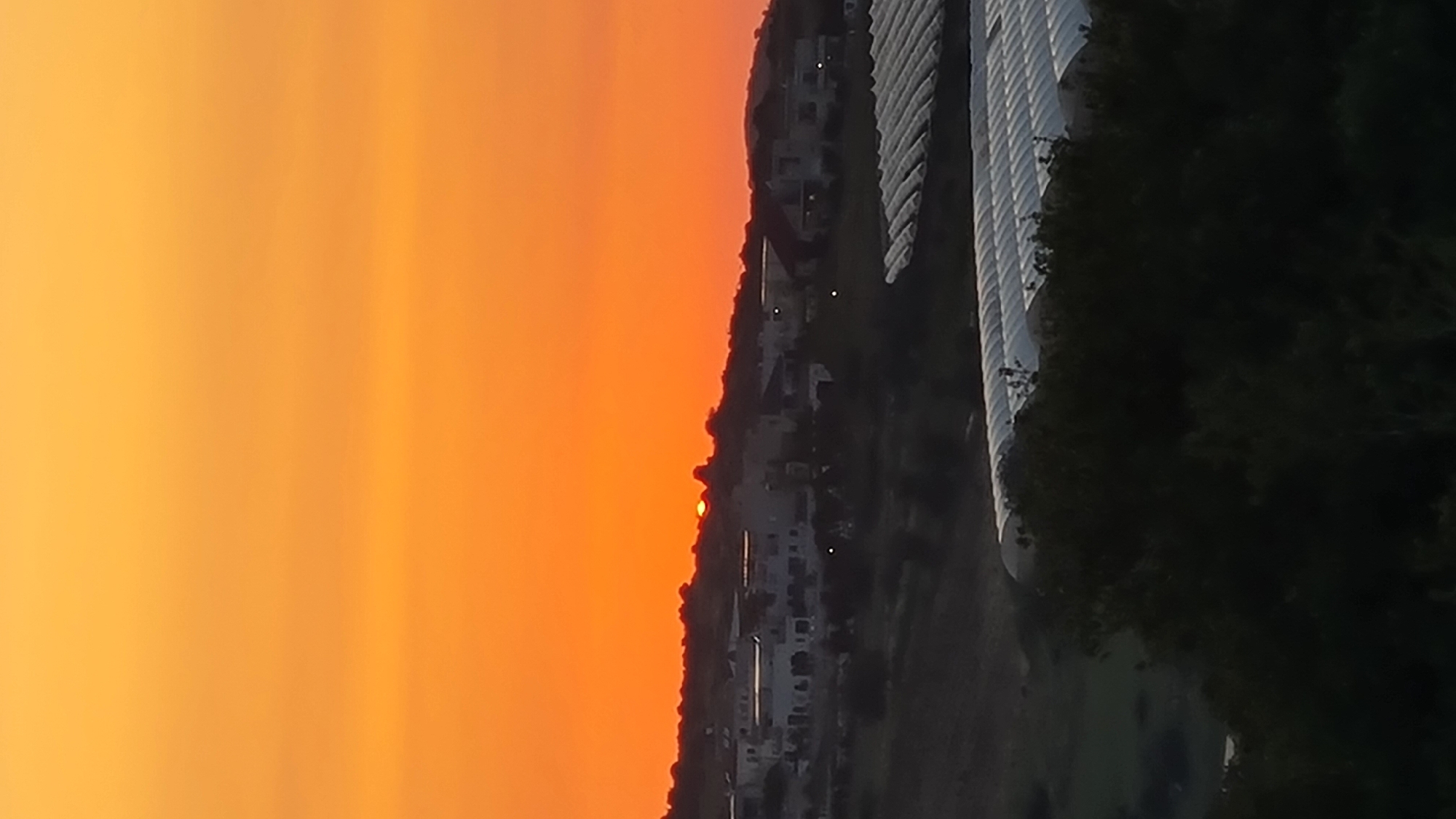 The image shows a sunset view in Lachish Regional Council, Israel. The sky is filled with orange and yellow hues. A settlement with white buildings is silhouetted against the skyline. To the right, dense dark green foliage obscures part of what appears to be a large white structure, potentially a building or complex. The sun is setting on the horizon.