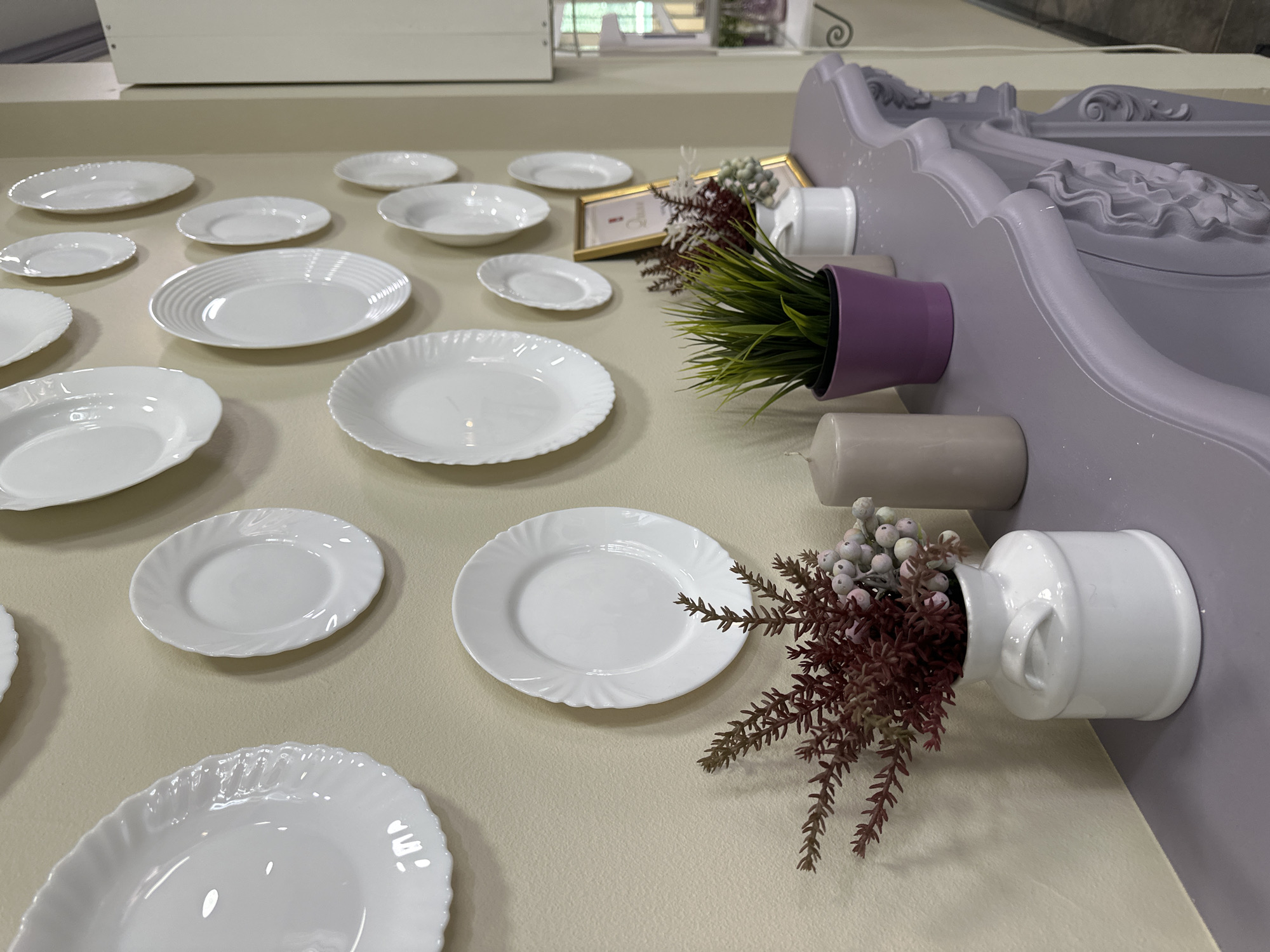 This is a medium-angle shot of a wall decorated with white plates. The plates are of various sizes and shapes, arranged in a somewhat irregular pattern. The wall is painted a neutral, light beige color.
On a decorative, light purple shelf or mantelpiece in the foreground, there are several objects. To the left, there is a white ceramic vase filled with a faux floral arrangement featuring reddish-brown branches and light blue berries. Next to the vase is a thick, grey candle. In the center of the shelf, there is a small purple pot containing a green faux plant. Behind the plant, a small gold-framed picture is leaning against the wall. Further to the right, there is a small white ceramic container.
The shelf itself has an ornate, carved design. The background is mostly the beige wall with the plates, but on the far right, a sliver of a white cabinet or wall is visible, along with some out-of-focus elements that appear to be part of a store or display.
The lighting appears to be artificial and slightly warm. There are no people visible in the image. The style suggests an interior decoration setting, possibly a home or a retail space.
The location is not specified in the image itself.