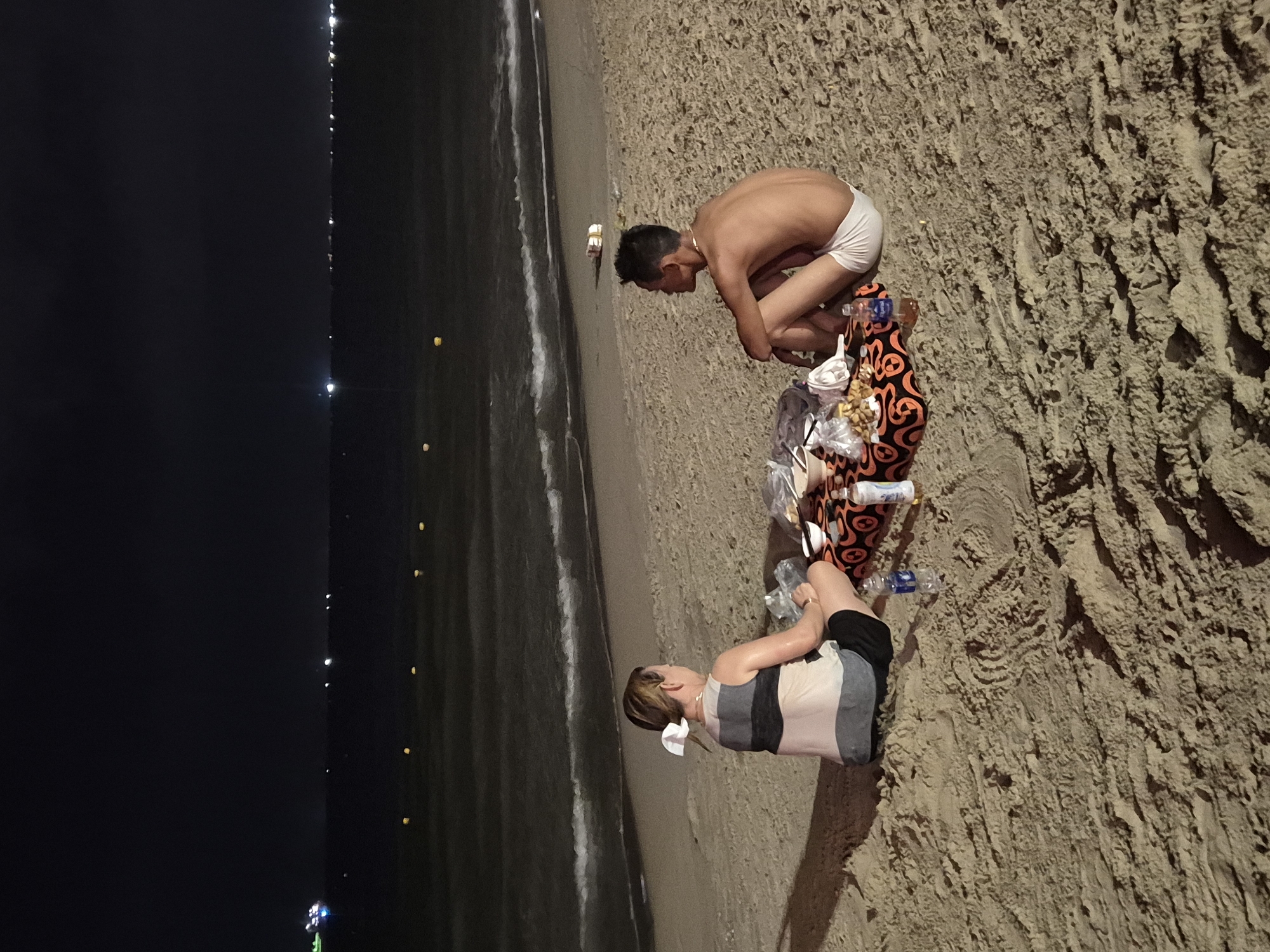 Two individuals, a woman and a man, are sitting on a sandy beach at night, facing each other, with a picnic spread between them. The woman, seen from behind, has her hair tied up with a white ribbon and is wearing a striped sleeveless top and dark shorts. The man, also seen from behind, is shirtless, wearing white shorts, and has his left arm resting on his knee. The picnic includes bags of food, bowls, and bottles of liquid. The beach extends towards a dark ocean with gentle waves and a distant horizon dotted with lights, possibly from boats or a city. The sky above is dark. The scene appears to be located on My Khe Beach in Đà Nẵng, Vietnam, a popular tourist destination known for its coastline.